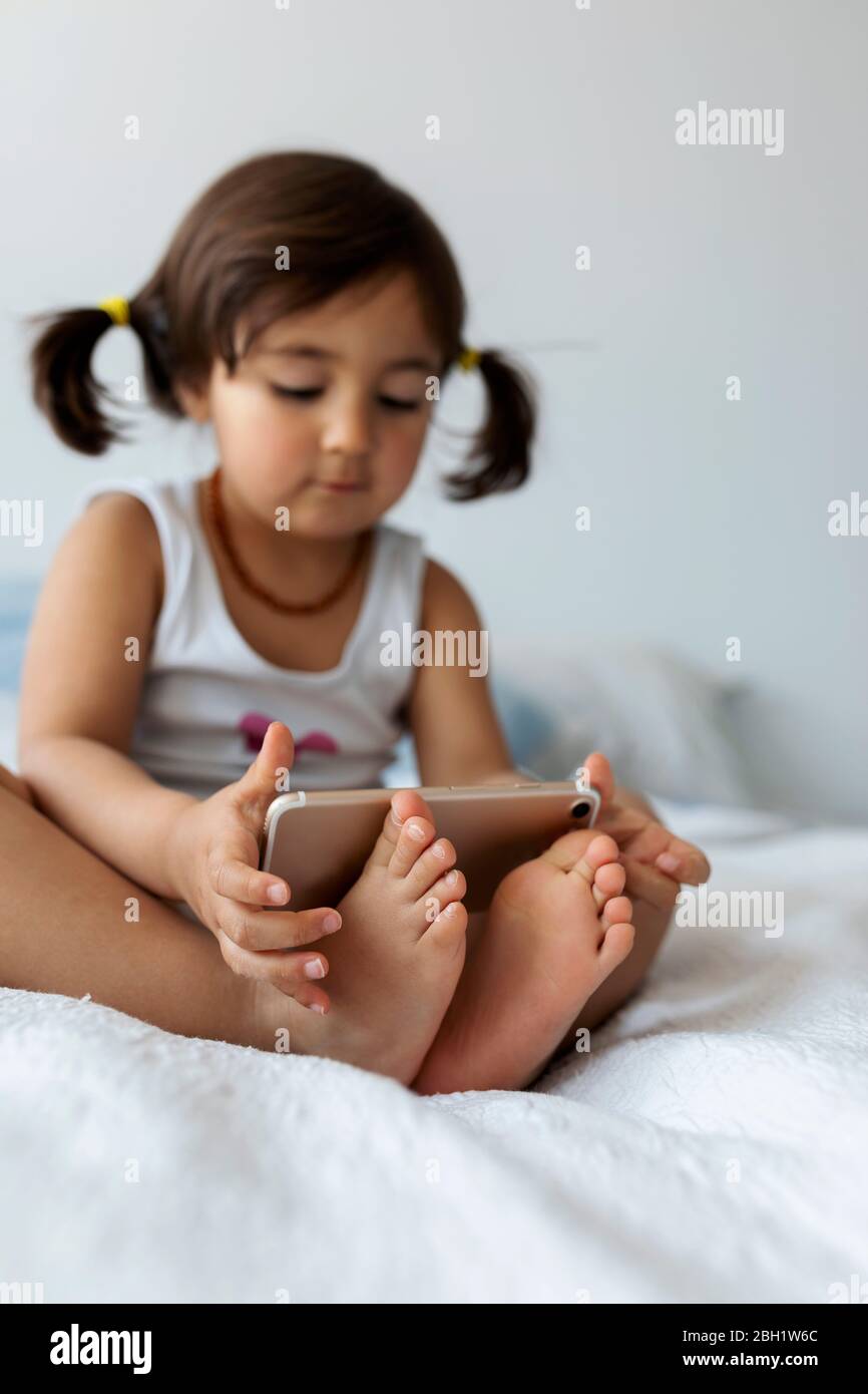 Petite fille assise sur le lit tenant un smartphone avec les pieds, gros plan Banque D'Images