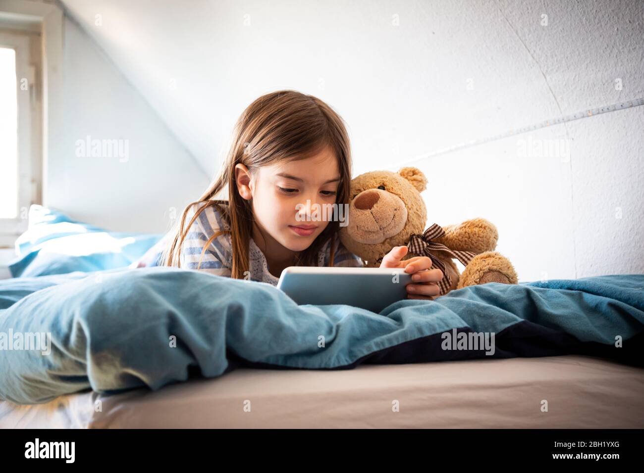 Portrait d'une fille couché sur un lit avec un ours en peluche à l'aide d'une tablette numérique Banque D'Images