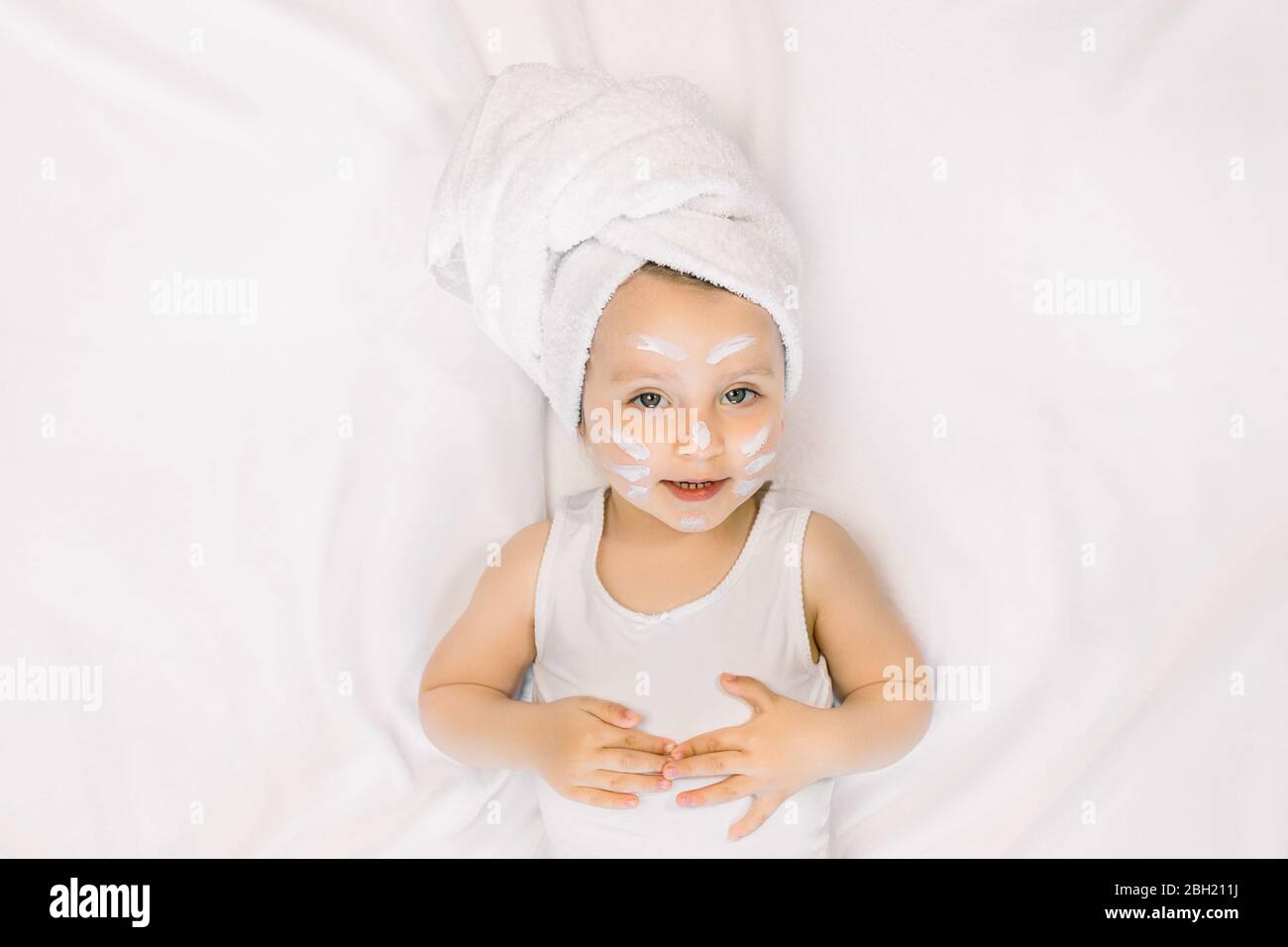 Petite Fille De Bebe Ayant Plaisir Apres Bain Avec Serviette Sur La Tete Et Creme De Visage Sur Les Joues Le Nez Et Le Front Couche Sur Fond Blanc Hygiene Du Bebe