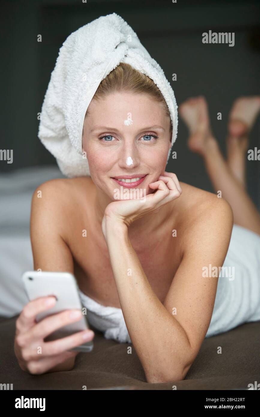 Portrait d'une femme magnifique avec tête enveloppée dans une serviette tenant un smartphone sur le lit Banque D'Images