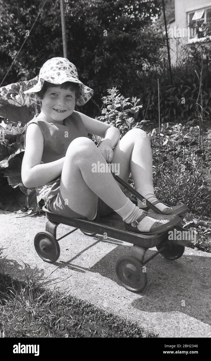 Années 1960, historique, une jeune fille portant un chapeau d'été et des sandales assis dans un petit chariot métallique à quatre roues sur un sentier dans un jardin à l'arrière, Angleterre, Royaume-Uni. Banque D'Images