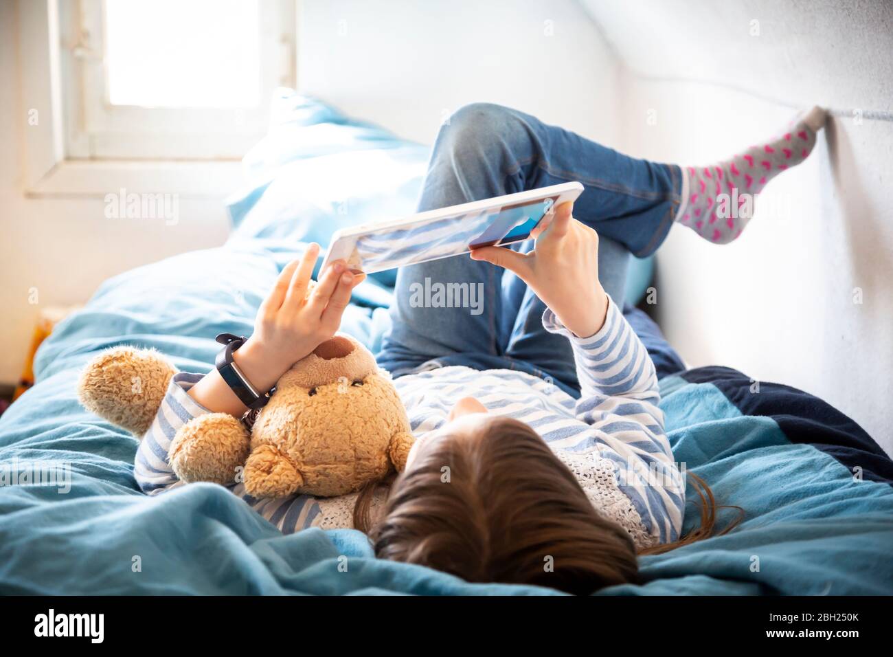 Petite fille couchée sur un lit avec un ours en peluche à l'aide d'une tablette numérique Banque D'Images