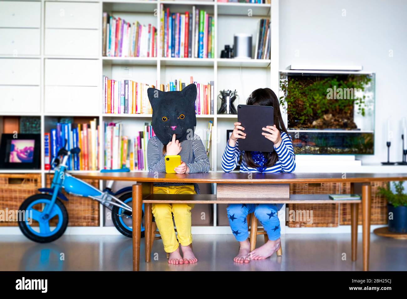 Deux filles assises à la table, se cachant derrière une tablette numérique et un masque pour chat Banque D'Images