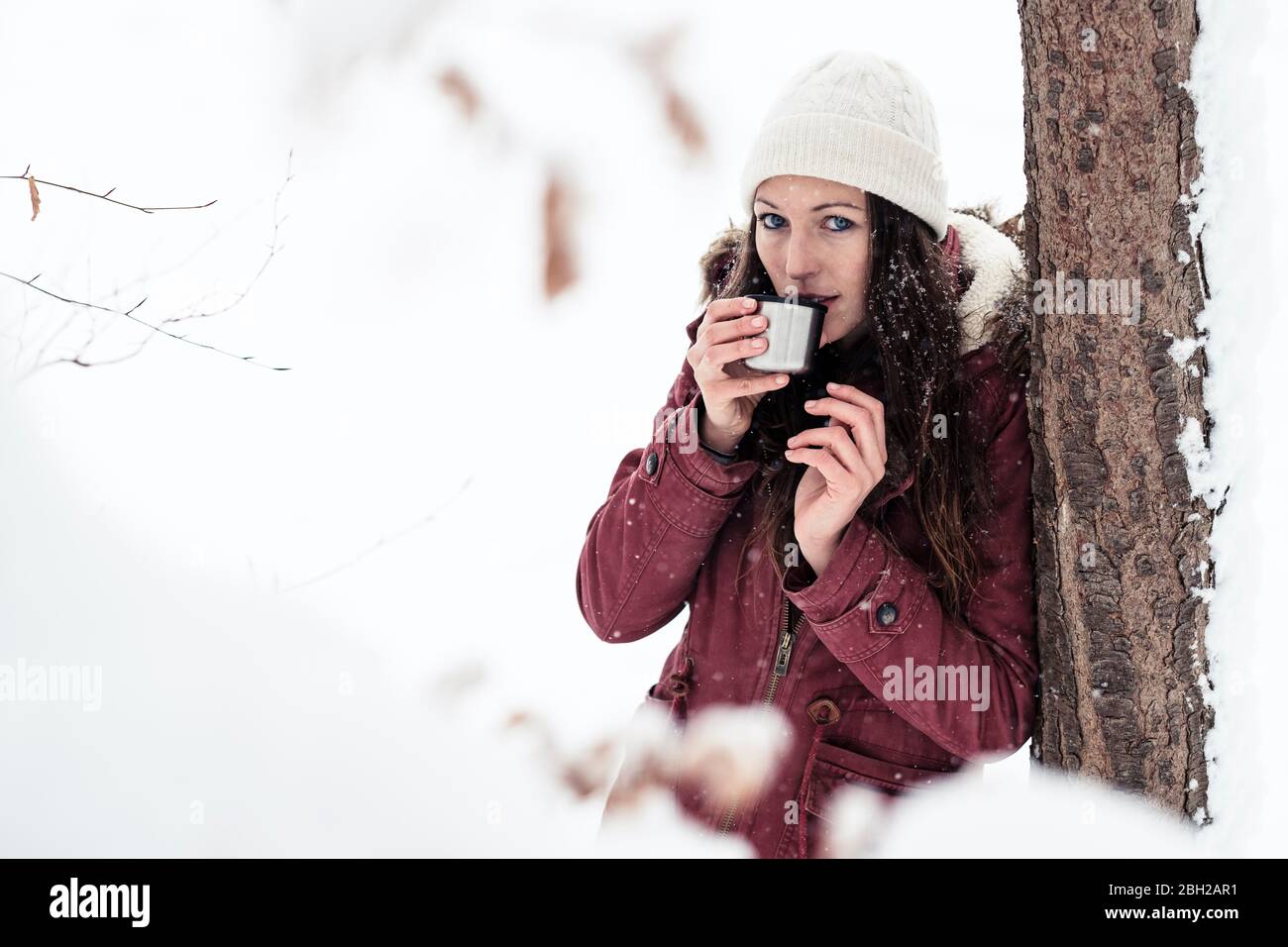 Portrait d'une jeune femme qui s'est penchée contre le tronc d'arbre en buvant une boisson chaude en hiver Banque D'Images