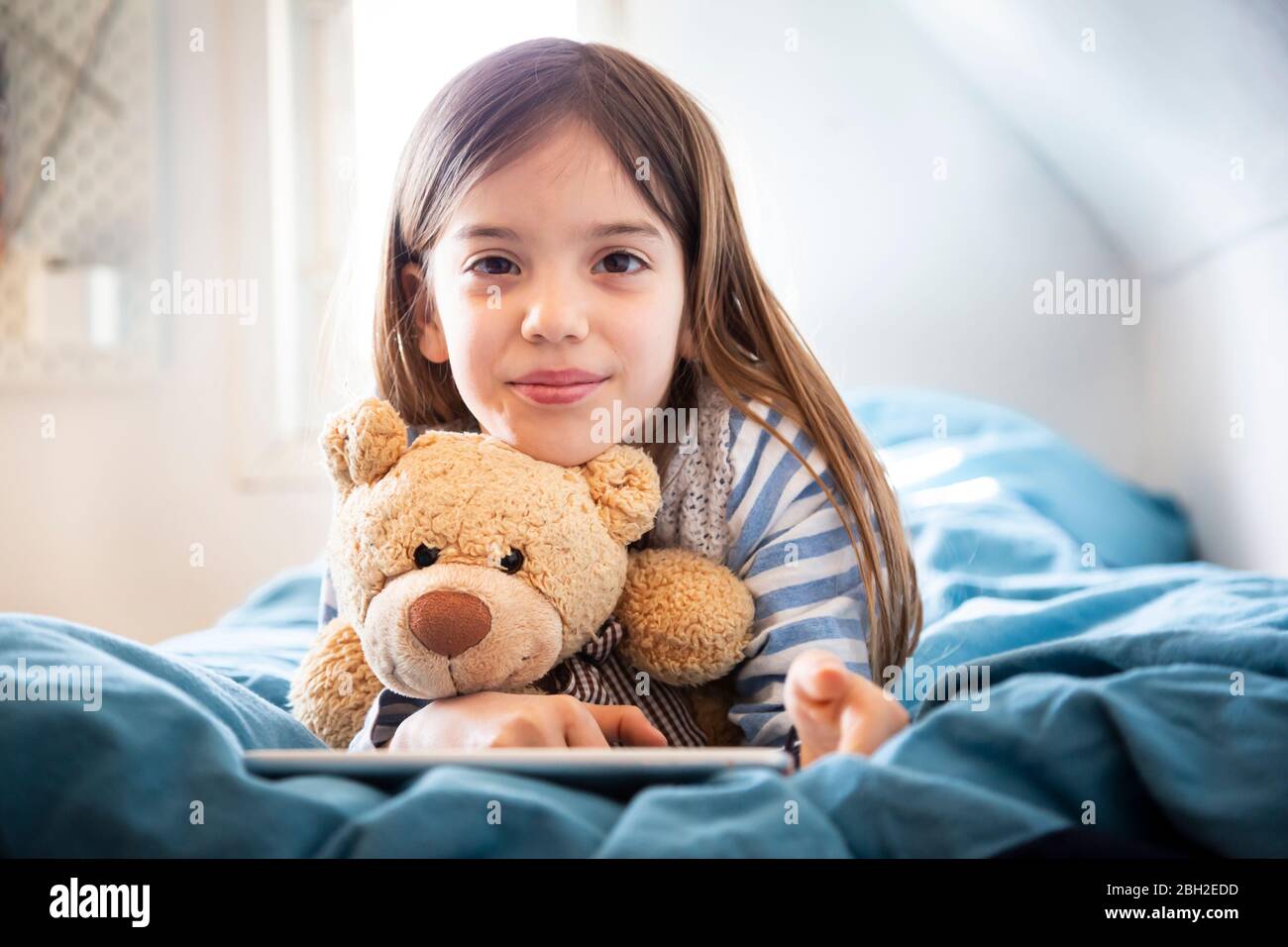 Portrait d'une fille souriante couché sur un lit avec un ours en peluche et une tablette numérique Banque D'Images