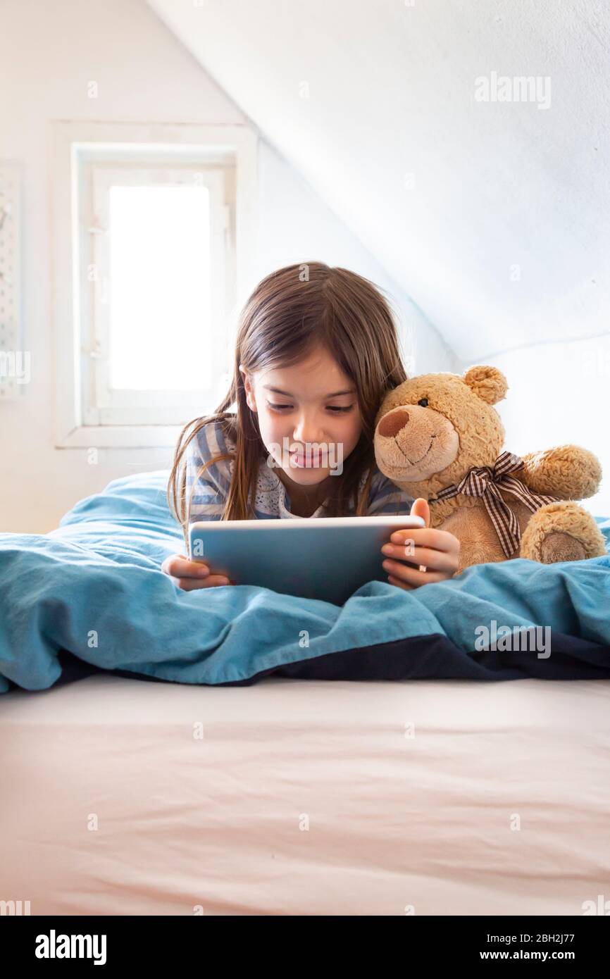 Portrait d'une jeune fille souriante couché sur un lit avec un ours en peluche à l'aide d'une tablette numérique Banque D'Images