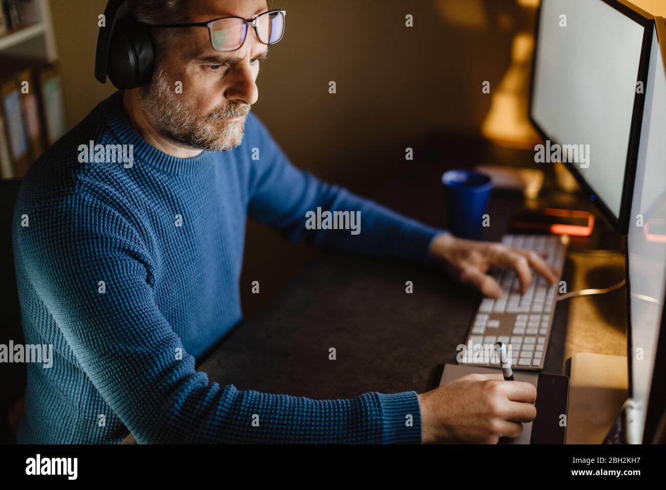 Homme mature avec casque assis au bureau à la maison travaillant avec une tablette graphique et un ordinateur Banque D'Images