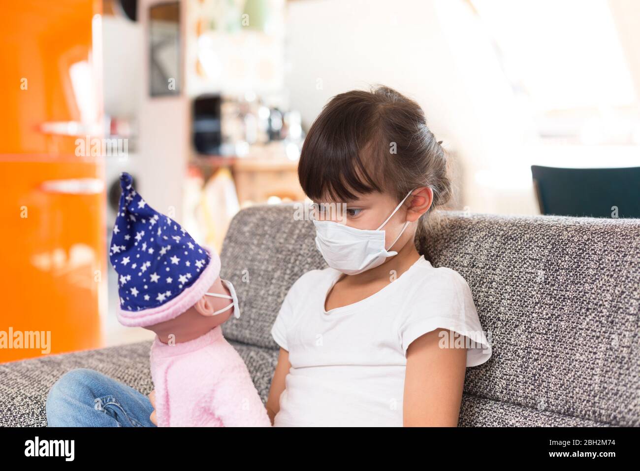 Petite fille avec masque protecteur assis sur le canapé à la maison jouant avec la poupée Banque D'Images