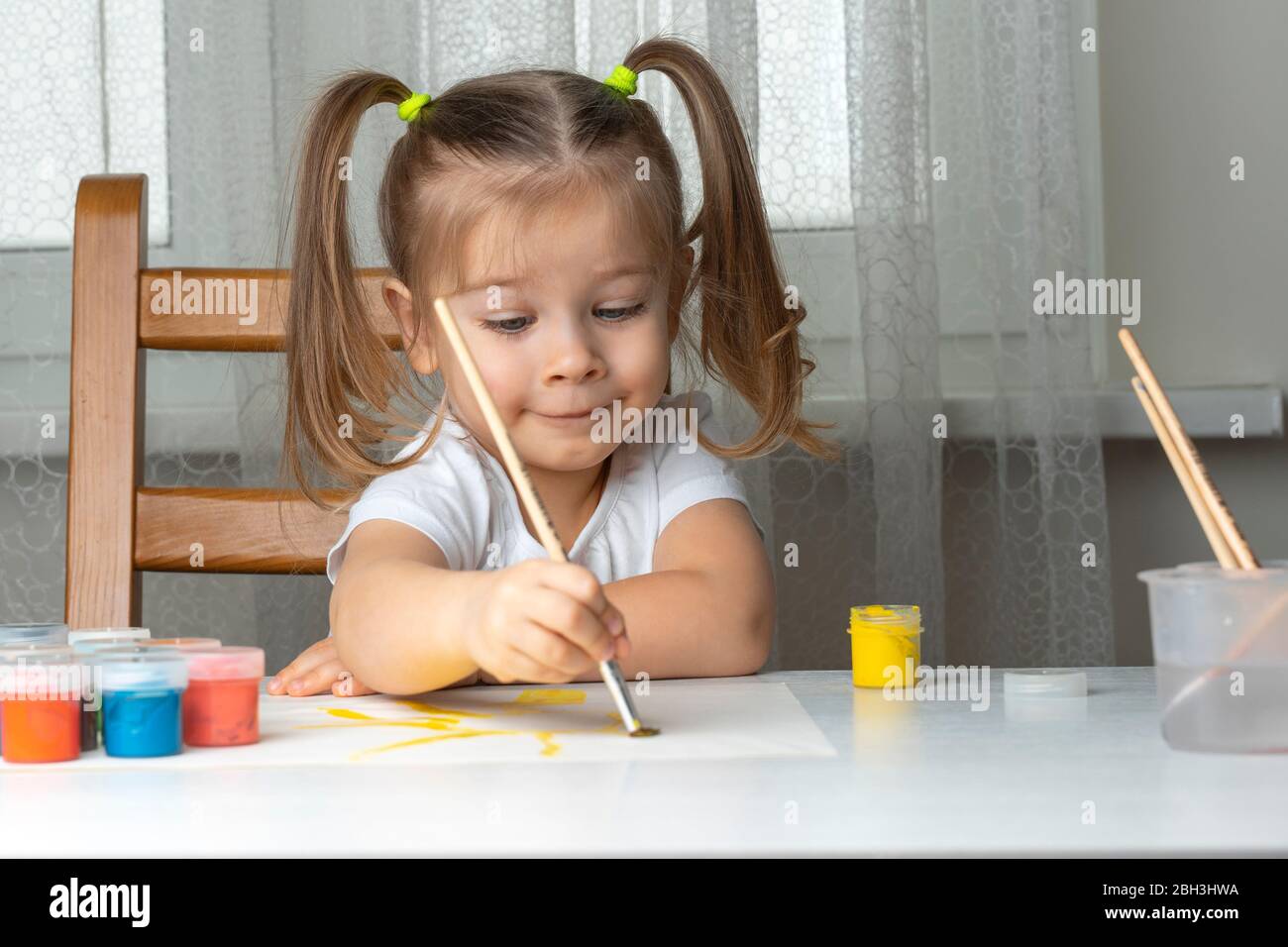 Petite belle fille de 3-4 ans avec deux queues de ponyles dans un T-shirt blanc peintures à la table et sourires. Mis en quarantaine la créativité des enfants en raison de cor Banque D'Images