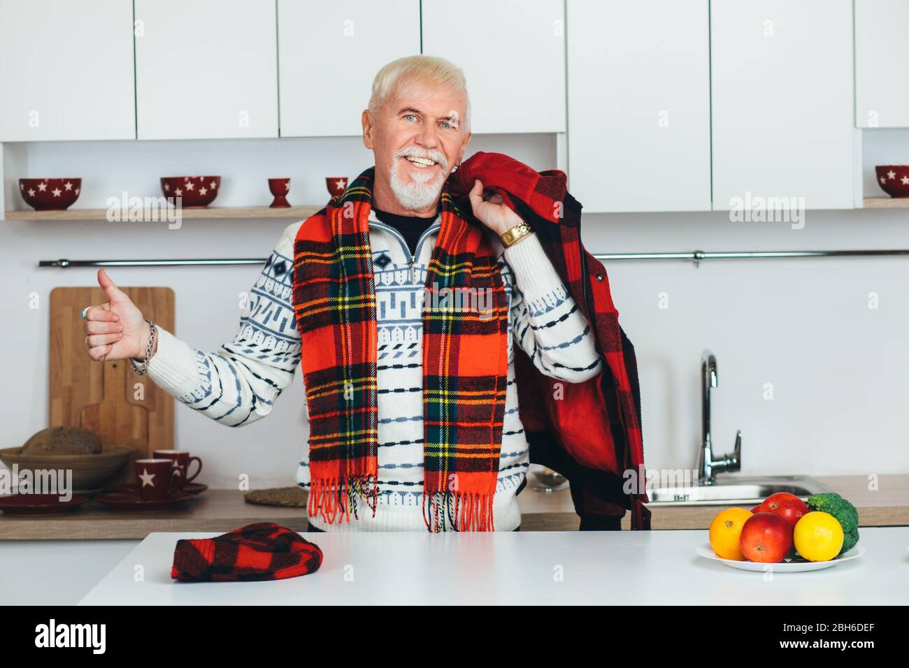 homme âgé debout en vêtements de dessus, écharpe à carreaux à la maison dans la cuisine, regardant l'appareil photo sourire et montrant des pouces vers le haut Banque D'Images