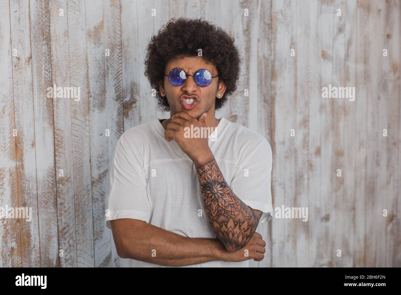 Être original ! L'homme aux cheveux bouclés positive, porter des lunettes avec motif cosmique , debout contre la paroi en bois Banque D'Images