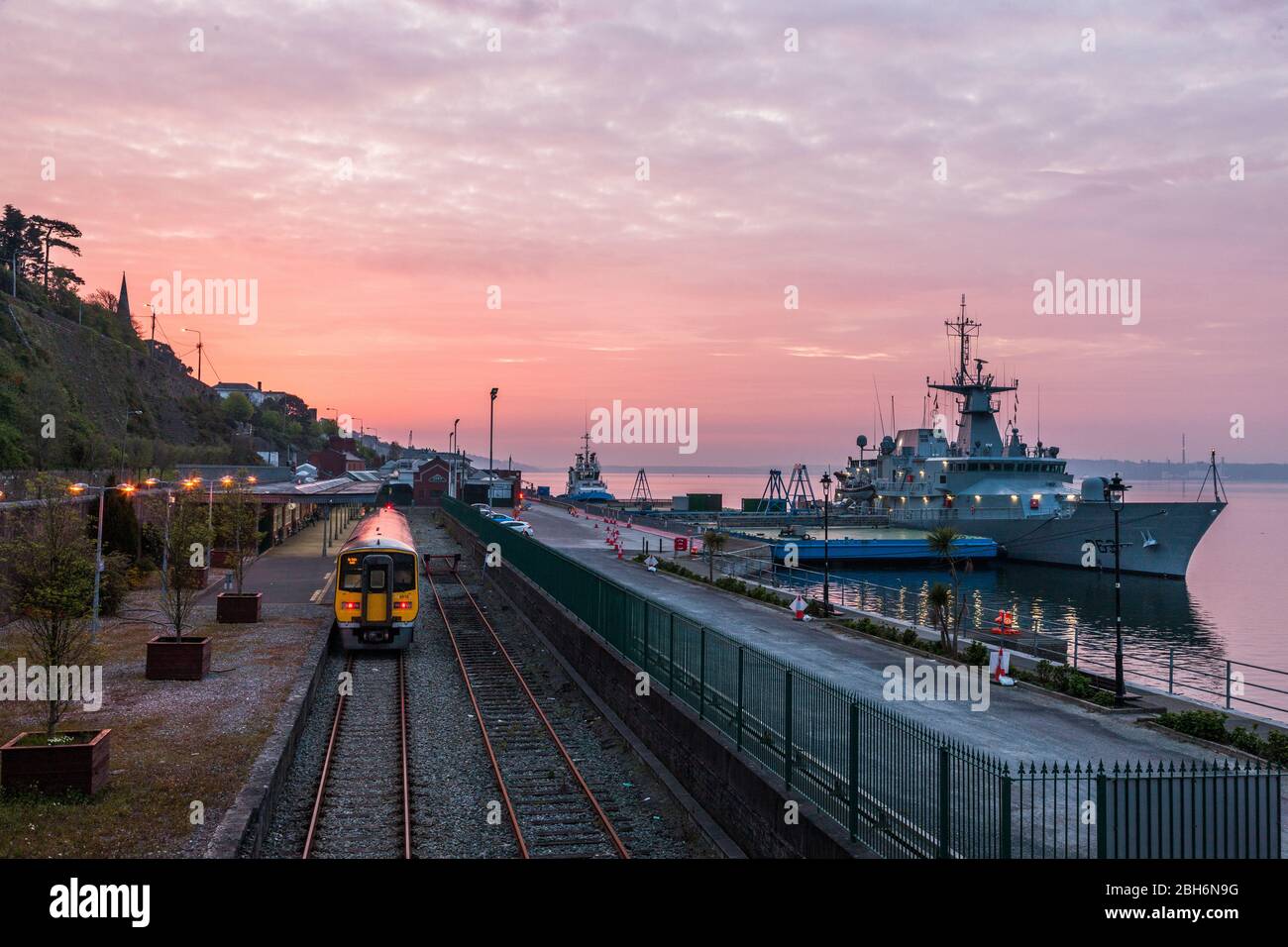 Cobh, Cork, Irlande. 24 avril 2020. Le train arrive tôt le matin à la gare, tandis que LE navire naval LÉ William Butler Yeats se trouve à la berth avant l'aube au quai des eaux profondes de Cobh, Co. Cork. Depuis le déclenchement de la pandémie de Coronavirus en Irlande, le service naval a apporté un soutien au service de santé en faisant des essais au Covid-19 à l'aide de leurs navires à Dublin, Galway et Cork. - crédit; David Creedon / Alay Live News Banque D'Images