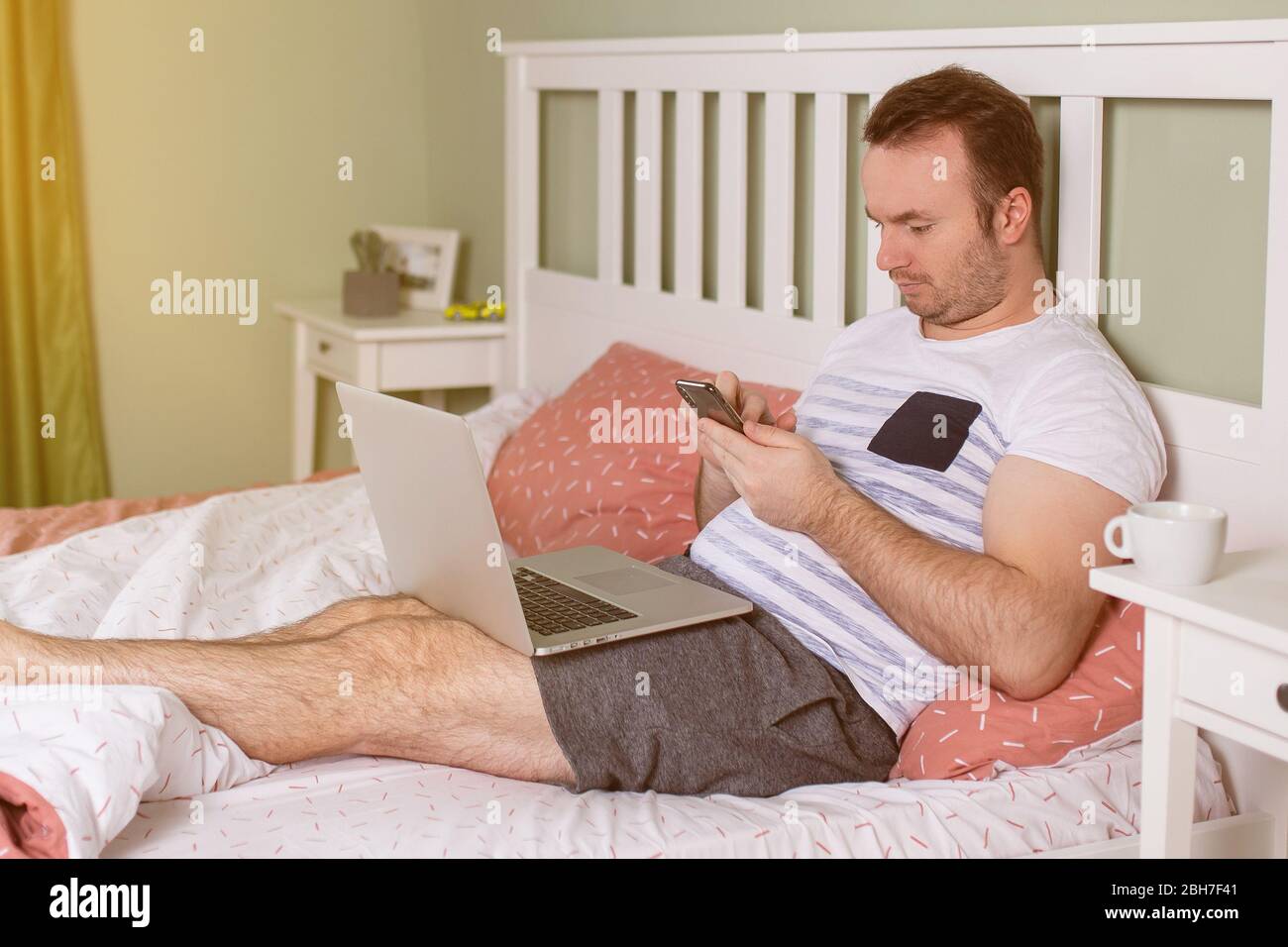 Beau jeune homme en pyjama à l'aide d'un ordinateur portable tout en allongé dans le lit le matin. Utilisation de son smartphone Banque D'Images