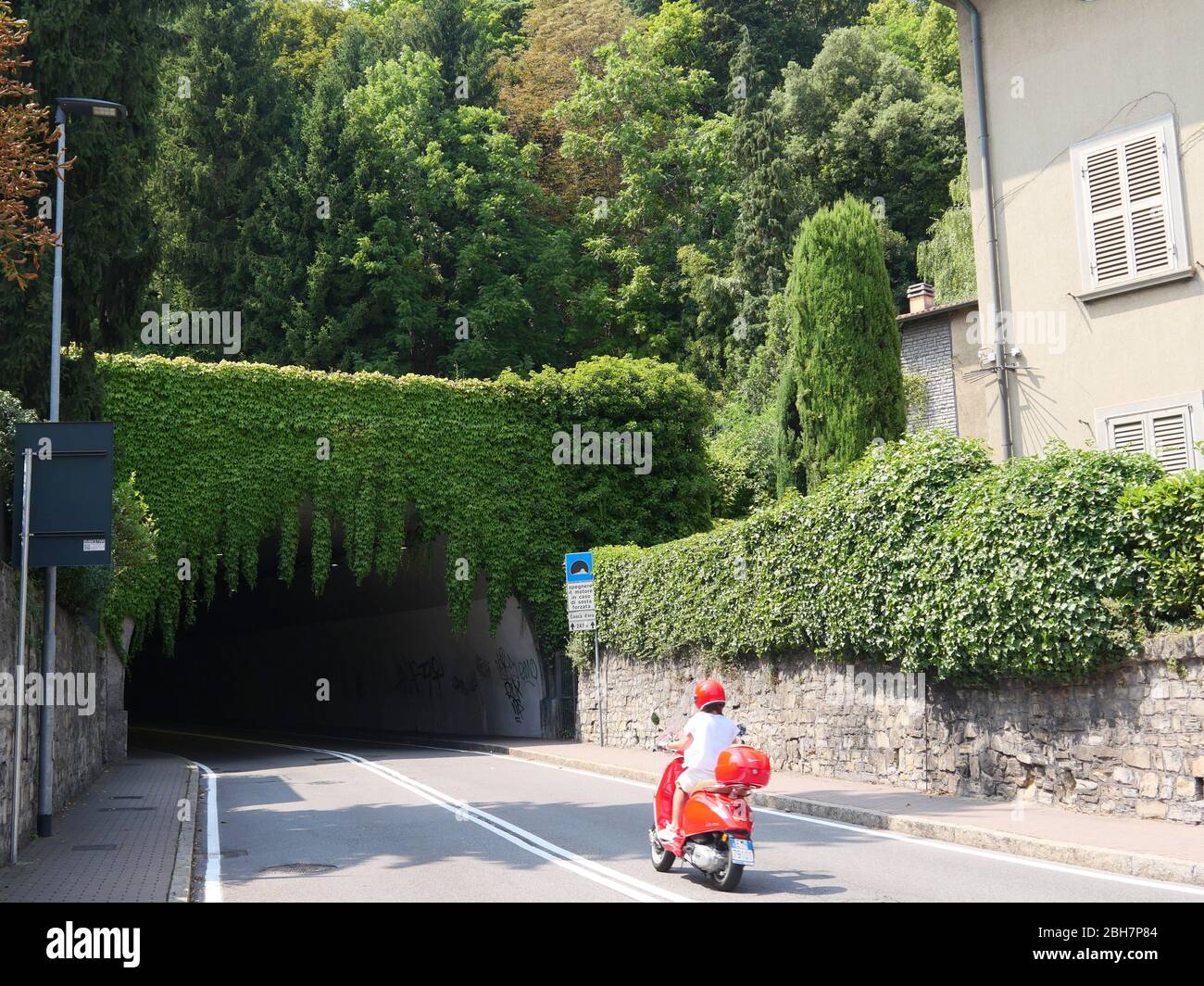 Bergame, Italie - 06 août 2019: Une fille conduisant un scooter Vespa rouge dans le beau tunnel vert couvert sombre Banque D'Images