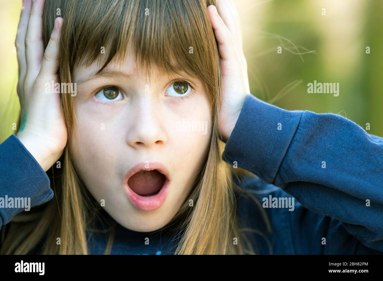 Portrait d'une jeune fille surprise tenant les mains à sa tête en plein air en été. Enfant de sexe féminin choqué pendant une journée chaude à l'extérieur. Banque D'Images