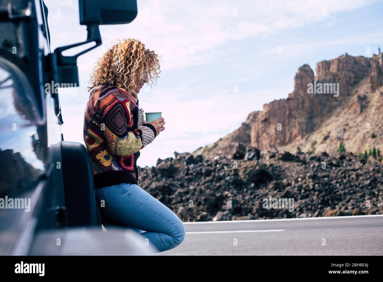 belle femme blanche d'âge moyen, blonde et indépendante, se reposer après un voyage avec sa voiture noire tout-terrain garée près de la rue. Montagnes et ciel en arrière-plan pour le concept d'aventure et les vacances alternatives Banque D'Images