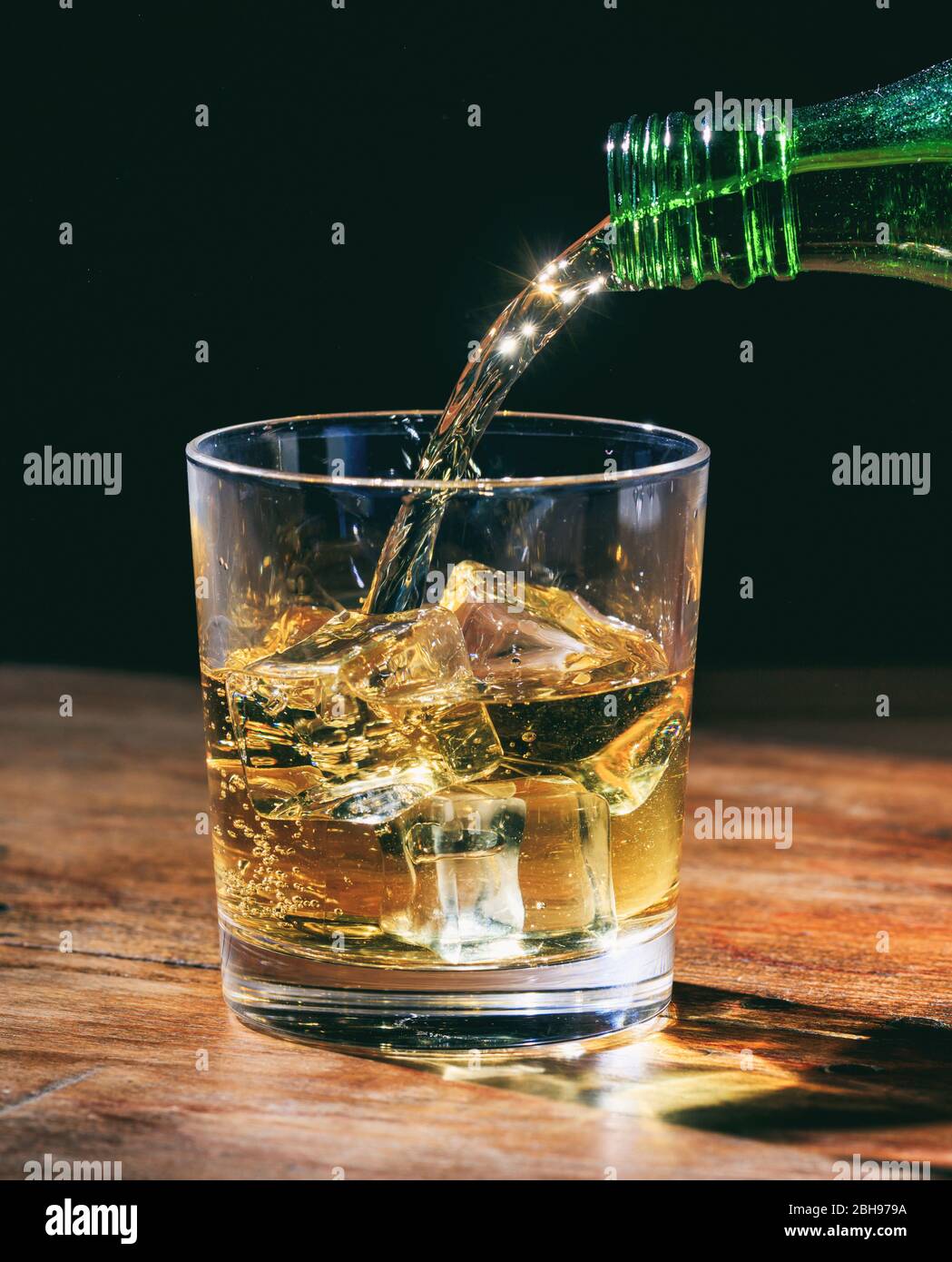 Bouteille verte qui verse du whisky sur les rochers, en verre cristal sur table en bois. Portrait vertical de la boisson alcoolisée avec des glaçons sur fond sombre. Banque D'Images