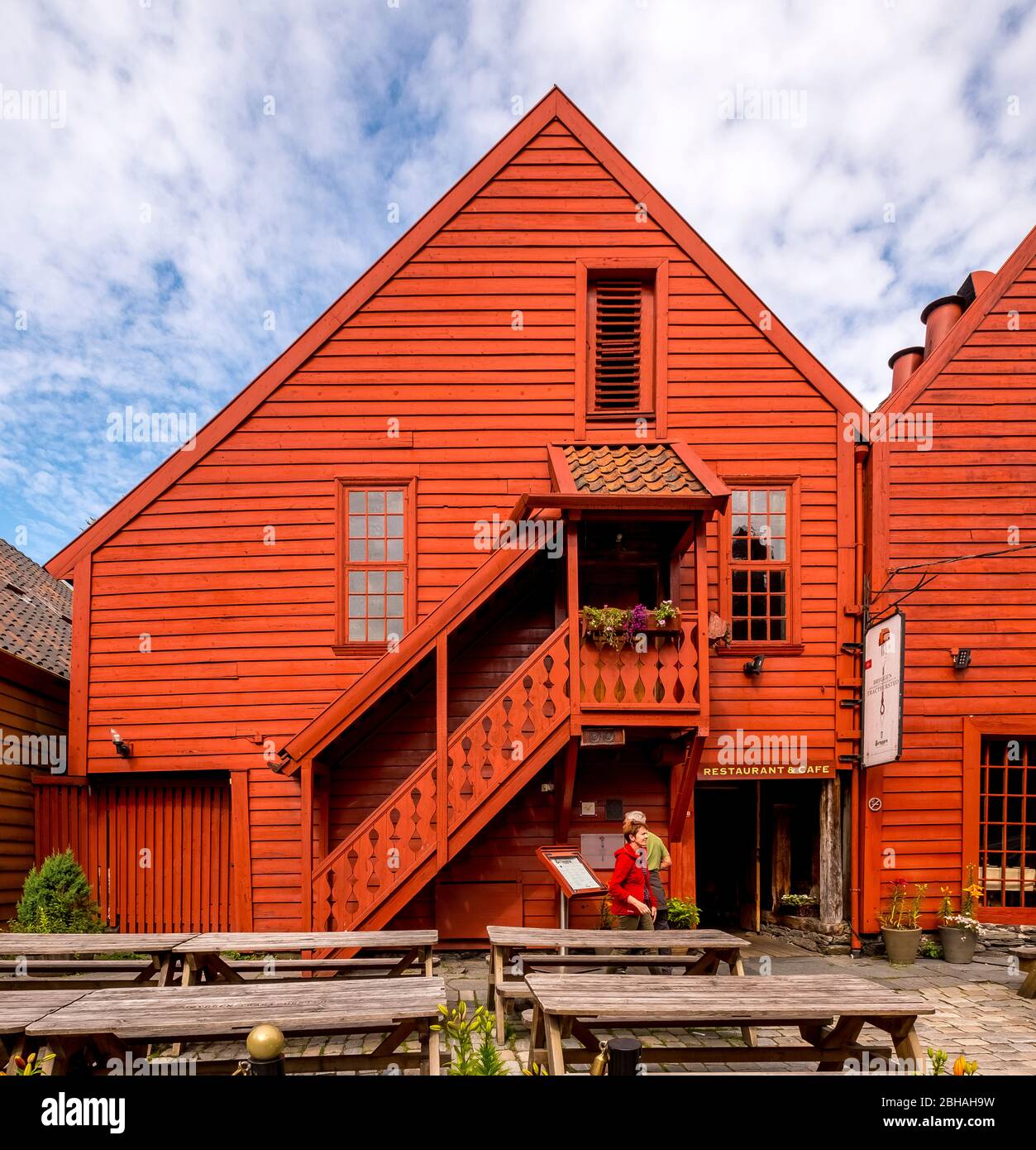 Les touristes marchent devant une maison en bois sculpturale avec escalier en bois, entourée d'une cour pavée sur les tables et les bancs, décorée avec des baignoires à fleurs, à Bryggen, Bergen, Hordaland, Norvège, Scandinavie, Europe Banque D'Images
