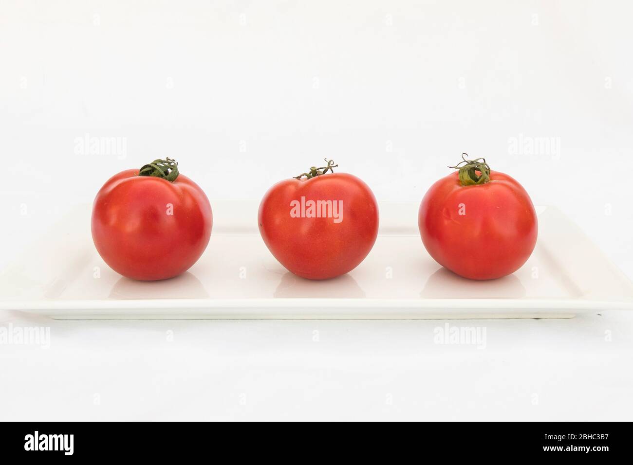 Trois tomates mûres, biologiques, rondes sur une plaque rectangulaire blanche avec un fond blanc Banque D'Images