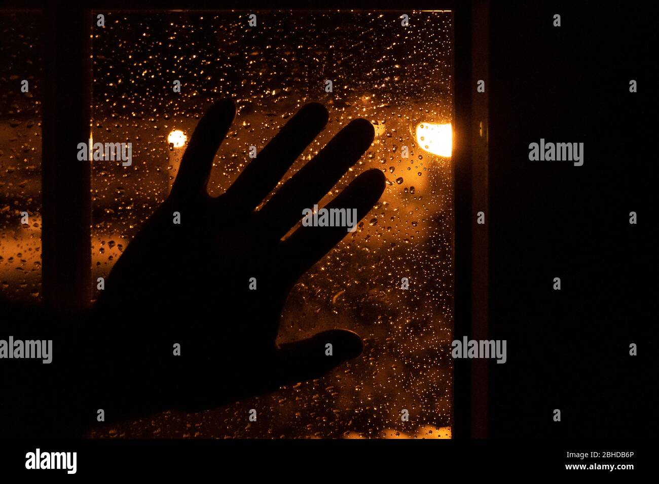 Silhouette d'une main reposant sur la fenêtre la nuit avec la lumière chaude des lampes de rue et avec des gouttes d'eau d'un jour pluvieux. Image triste de mel Banque D'Images