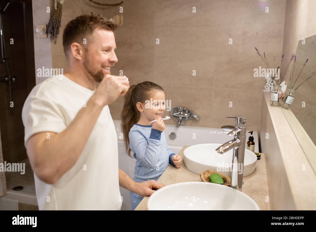 Joyeux jeune père barbu et sa petite fille gaie qui tient des brosses à dents tout en se tenant dans la salle de bains Banque D'Images
