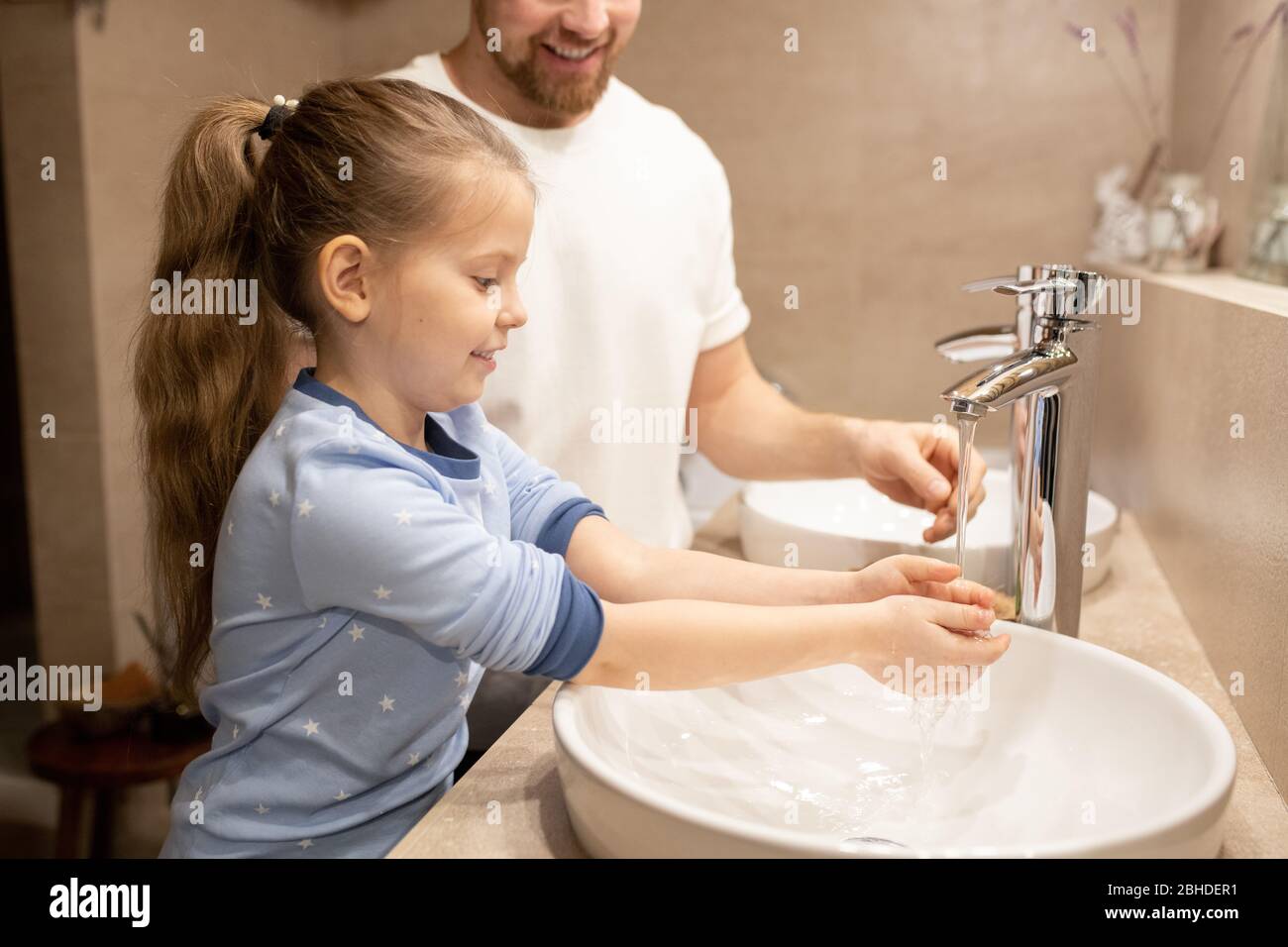 Bonne fille en pyjama bleu se lavant les mains sur l'évier le matin sur fond de son père debout près de dans la salle de bains Banque D'Images