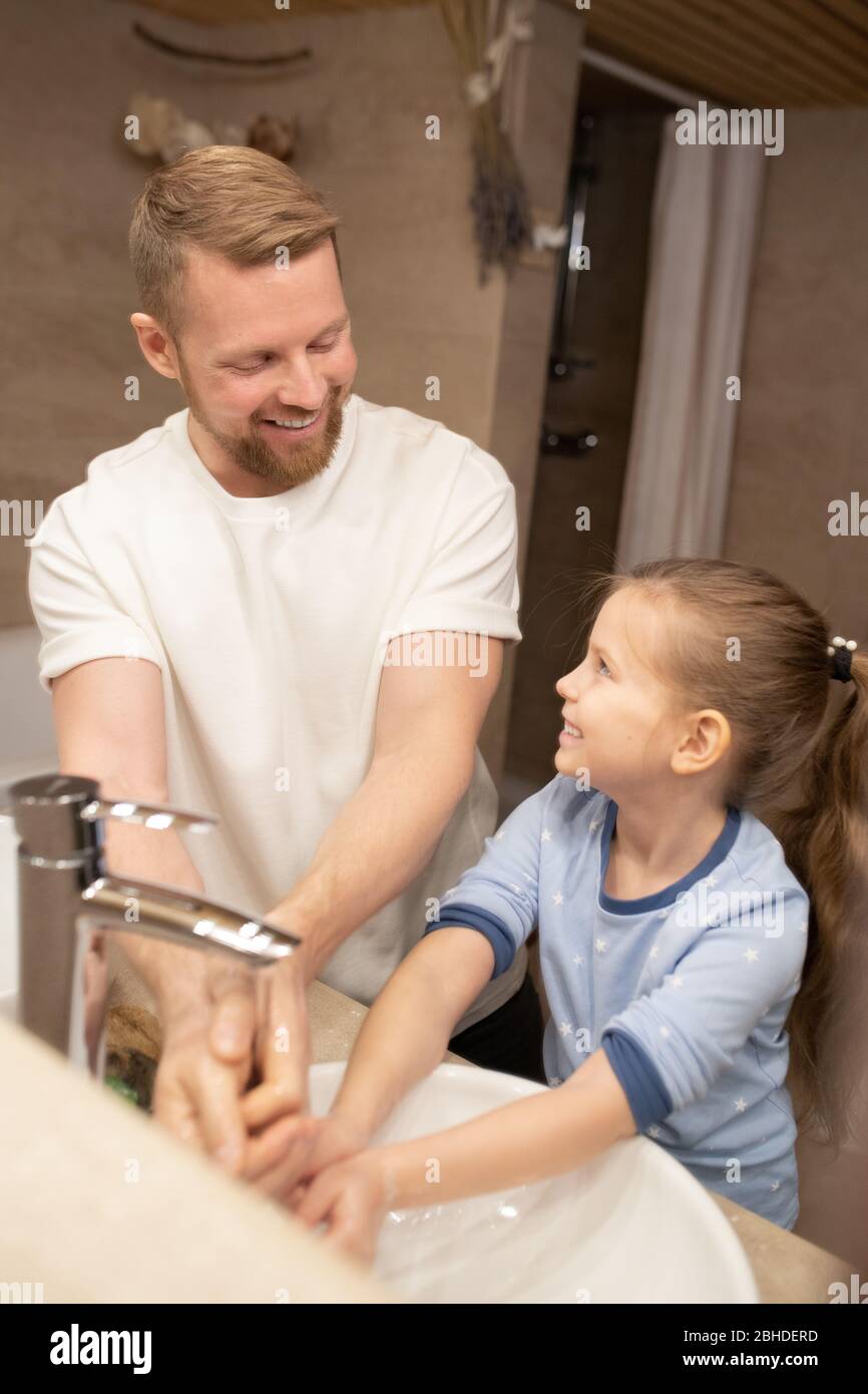 Jeune homme gai et sa petite fille adorable en pyjama regardant l'un l'autre tout en se tenant dans la salle de bains et se lavant les mains Banque D'Images