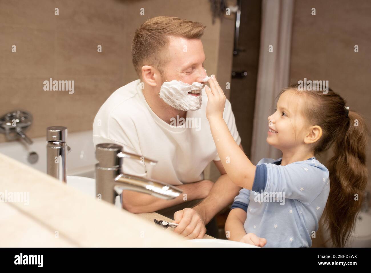 Joyeux adorable petite fille en pyjama touchant son nez de son père avec de la mousse à raser sur le visage et s'amuser dans la salle de bains le matin Banque D'Images