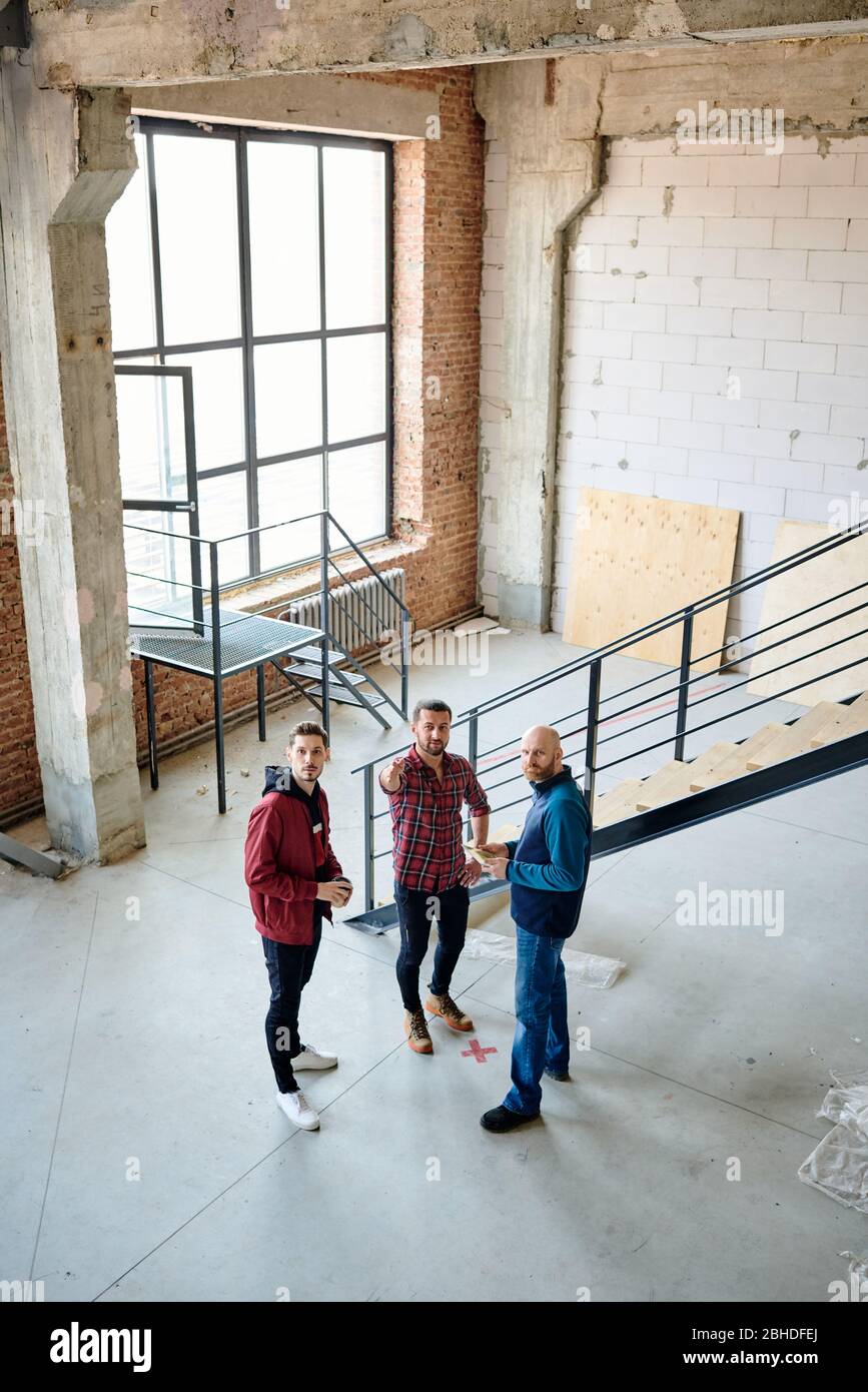 Groupe de jeunes ingénieurs ou entrepreneurs debout par escalier tout en discutant de la qualité du travail des constructeurs dans la construction inachevée Banque D'Images