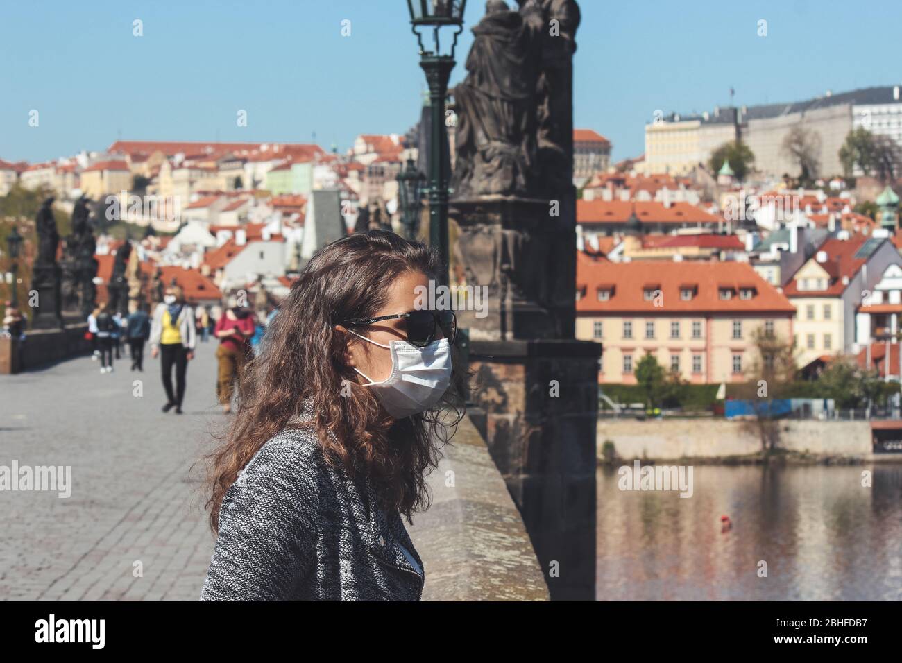 Jeune femme avec lunettes de soleil et masque médical photographié sur le pont Charles à Prague, République tchèque. Les gens sont floutés et la vieille ville en arrière-plan. Tourisme pendant le coronavirus. COVID-19. Banque D'Images