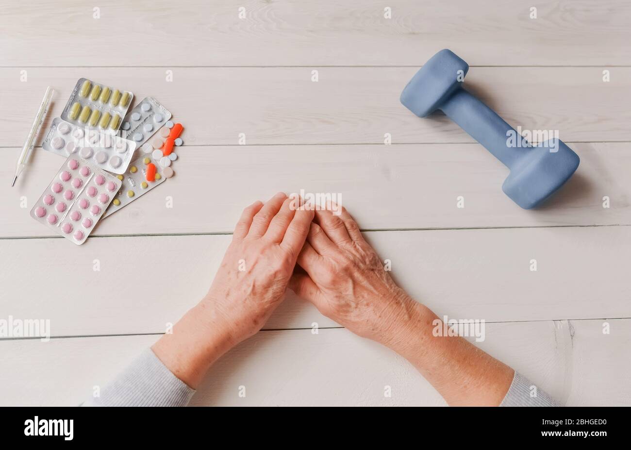Mains senior avec pilules, médicaments sur table, haltère. Femme froissée les mains, thermomètre, comprimés colorés et haltère, fond en bois. Soins de santé Banque D'Images