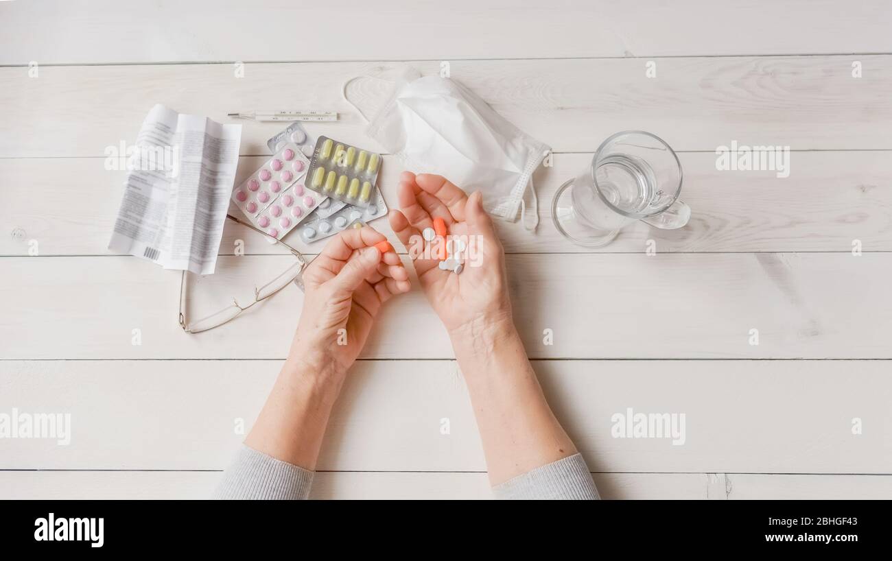 Mains, pilules et médicaments sur table, verre d'eau. Mains froissées de la vieille femme tenant des comprimés colorés, masque, lunettes, ordonnance, thermomètre Banque D'Images