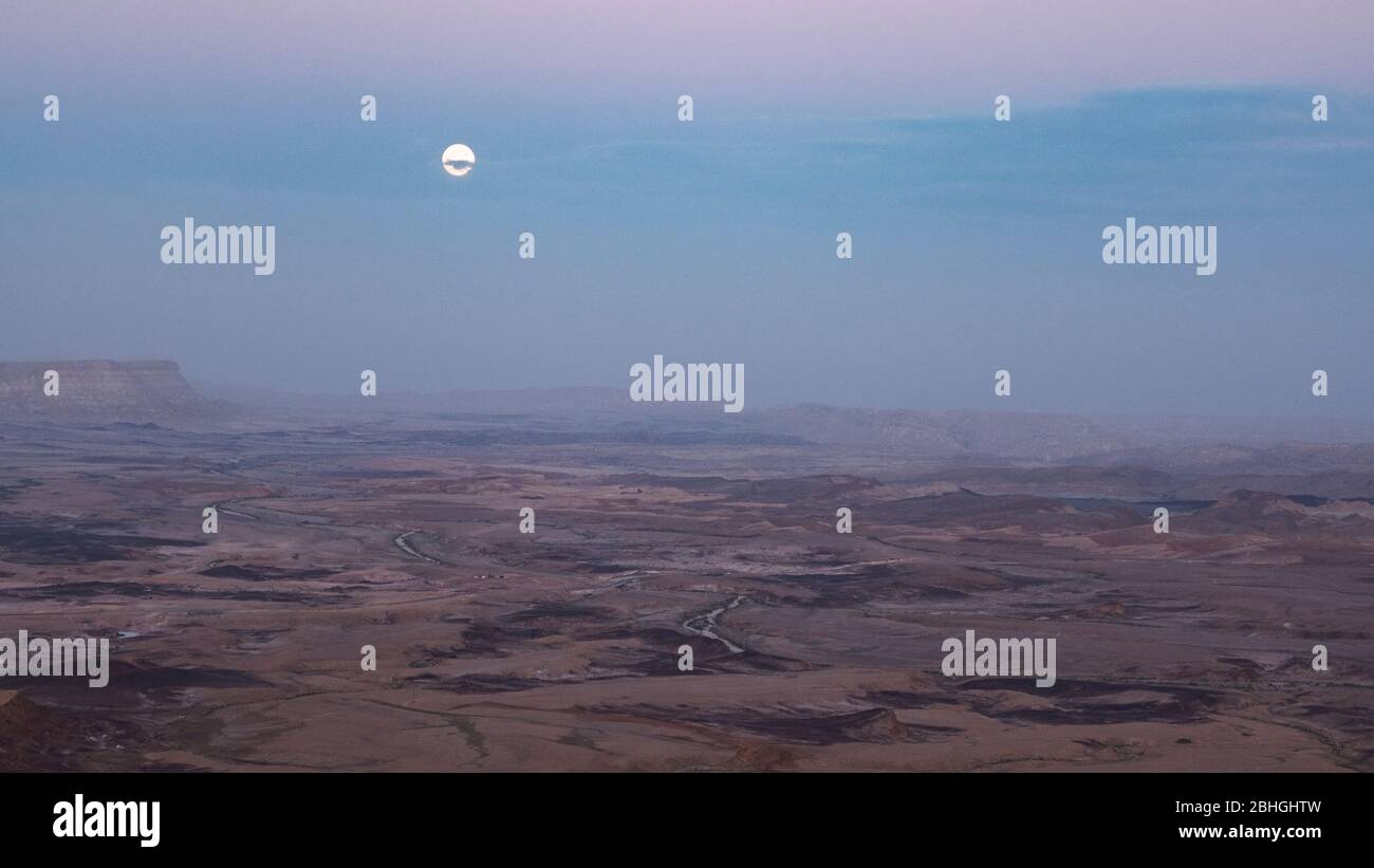 la pleine lune se fait piquer à travers des nuages au-dessus du cratère de makhtesh ramon en israël juste après le coucher du soleil à mitzpe ramon en israël Banque D'Images