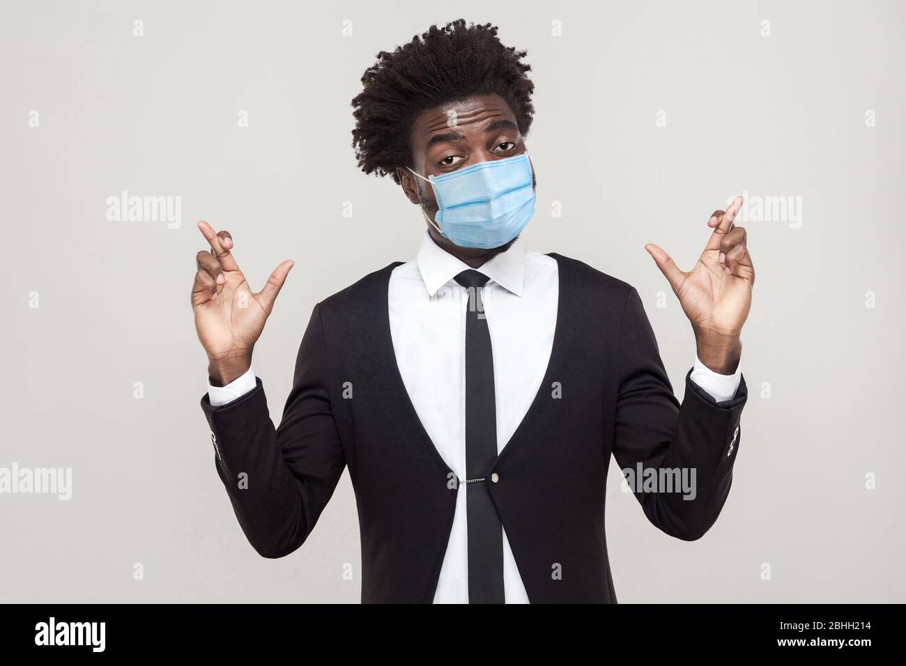 Plein d'espoir ou de vœu. Portrait de l'inquiétude jeune homme de main de travail portant costume noir avec masque médical chirurgical debout avec des doigts croisés et espoir. Banque D'Images