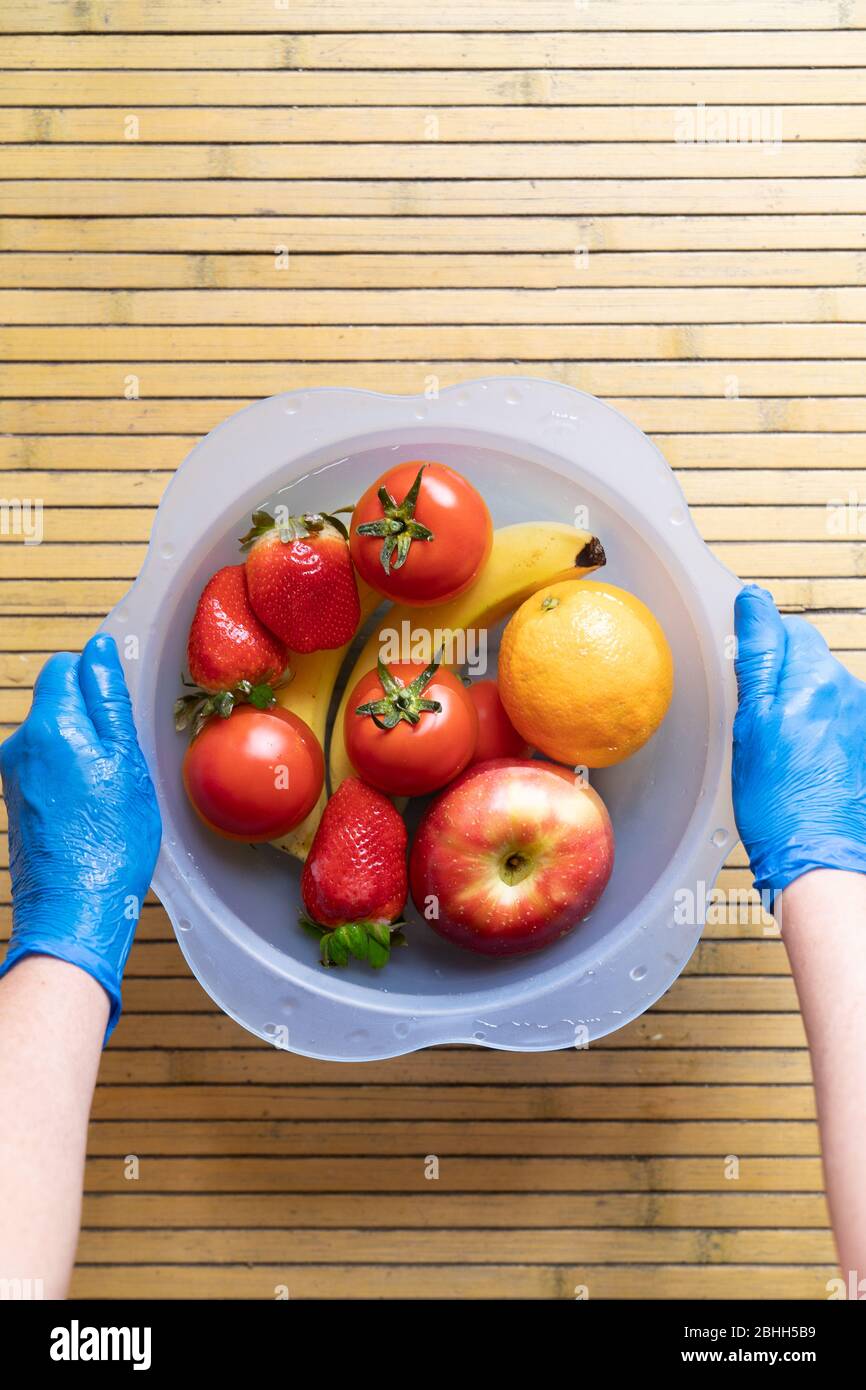 Mains avec gants en latex bleu remuant un bol bleu avec différents fruits frais et propres sur une base en bois. Bananes, tomates, pommes, fraises et Banque D'Images