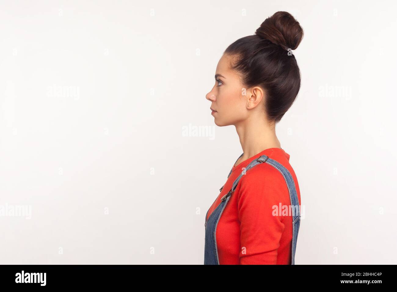 Profil d'une fille élégante et séduisante avec un pain pour cheveux dans des salopettes en denim debout avec un visage sérieux, expression pensive calme, regardant le spac de copie latérale Banque D'Images