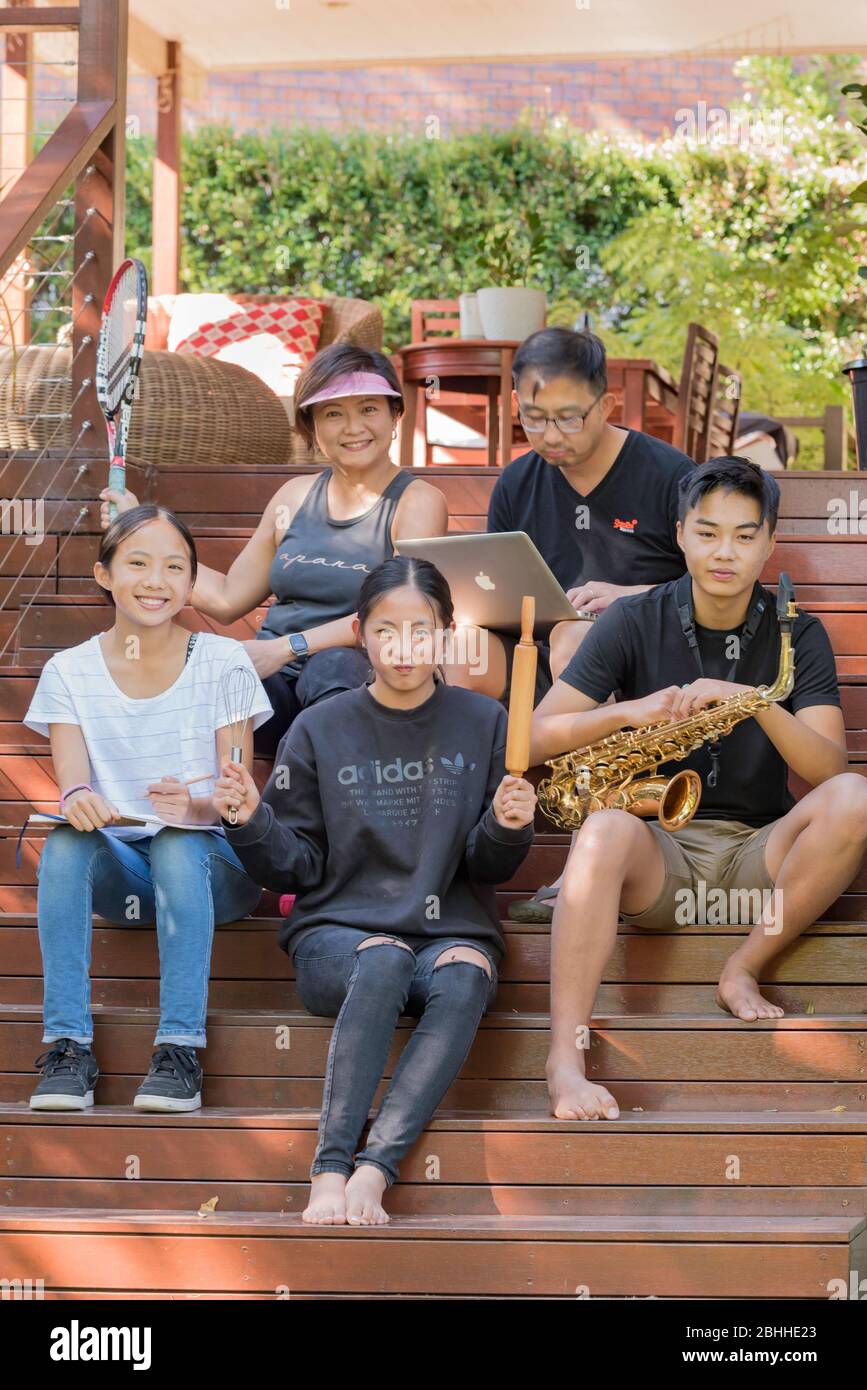 Une famille malaisienne australienne heureuse s'assoit sur les marches avant de leur maison de Sydney pendant l'épidémie de Covid-19, chacune montrant son passe-temps préféré. Banque D'Images