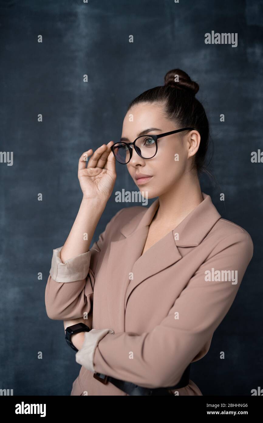 Jolie jeune femme réfléchie dans une veste élégante, se tenant contre le tableau noir et ajustant les lunettes tout en regardant de côté Banque D'Images