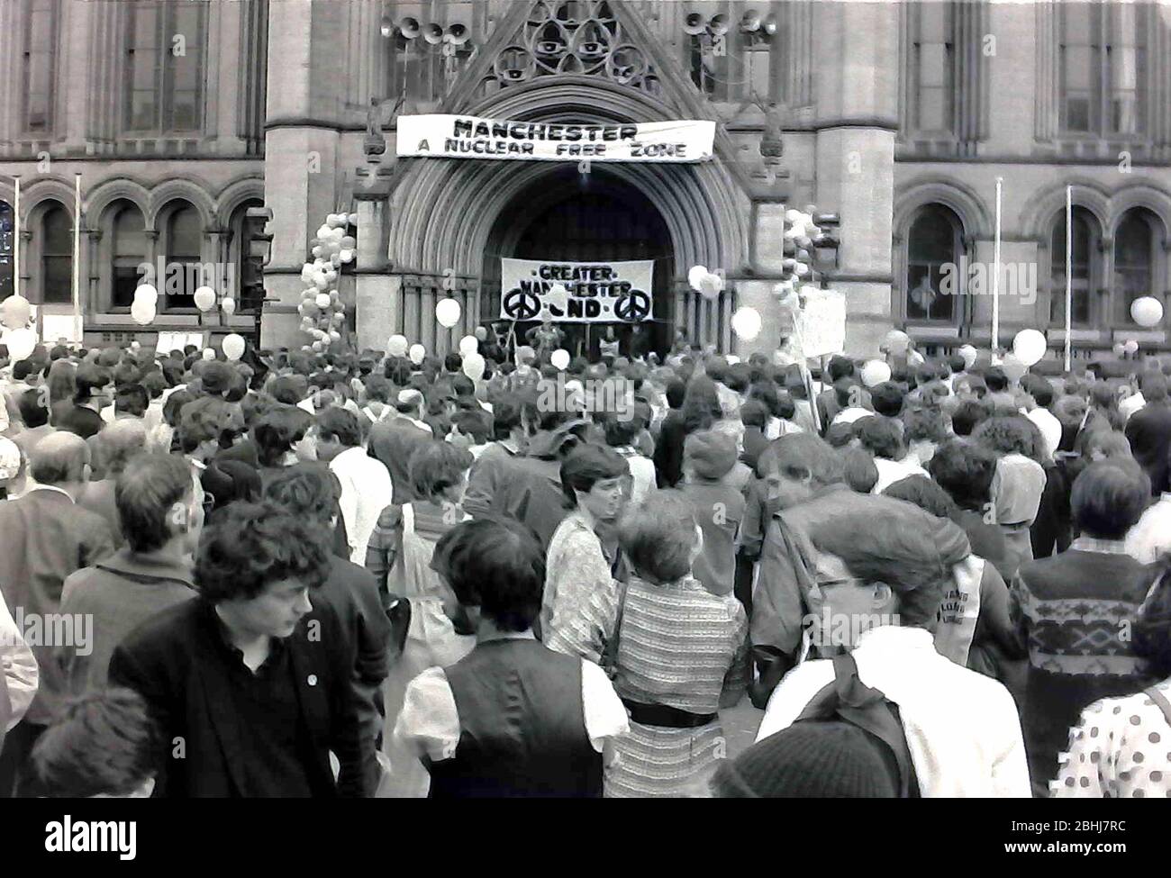 Les gens écoutent un orateur lors d'un rassemblement de campagne pour le désarmement nucléaire à Albert Square, à l'extérieur de l'hôtel de ville de Manchester, Grand Manchester, Angleterre, Royaume-Uni en 1983. Le Conseil municipal de Manchester a suspendu sa bannière « zone nucléaire libre de Manchester » au-dessus de l'entrée pour soutenir le pays. Banque D'Images