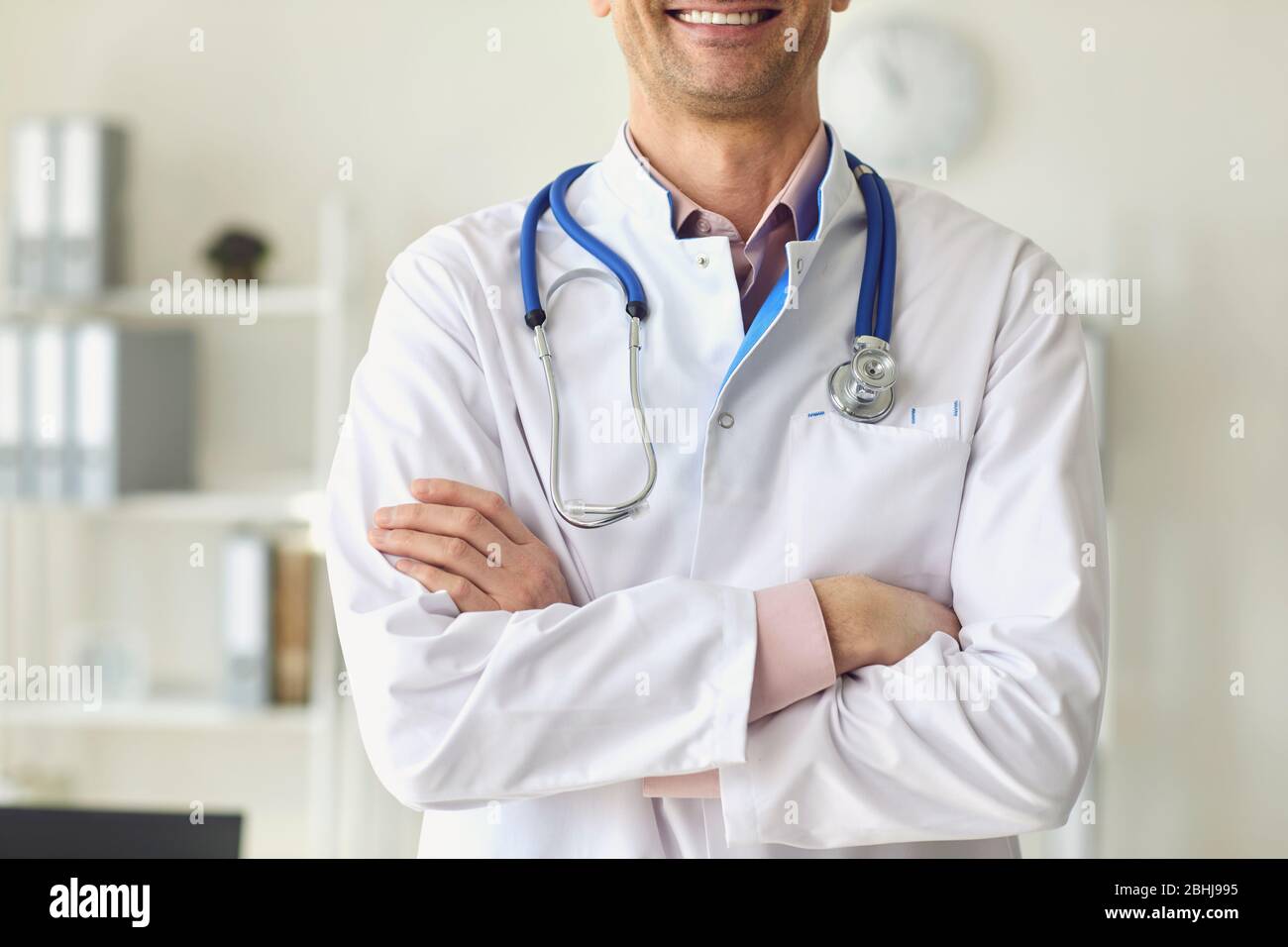 Un médecin thérapeute positif dans un manteau blanc a croisé des bras debout dans un bureau blanc d'une clinique médicale. Banque D'Images