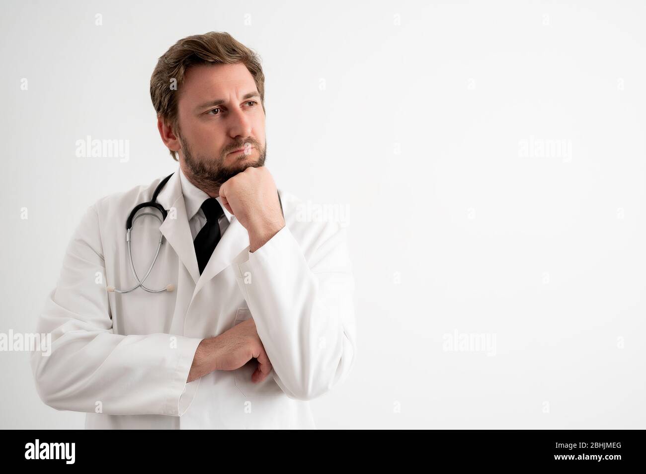 Portrait d'un médecin mâle avec stéthoscope en uniforme médical tenant la main sous son menton, ayant douté poser sur un fond blanc isolé Banque D'Images