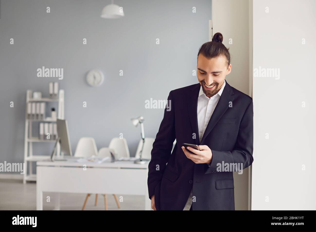Un homme d'affaires dans une veste noire debout lit un message dans un téléphone portable dans un bureau blanc. Banque D'Images