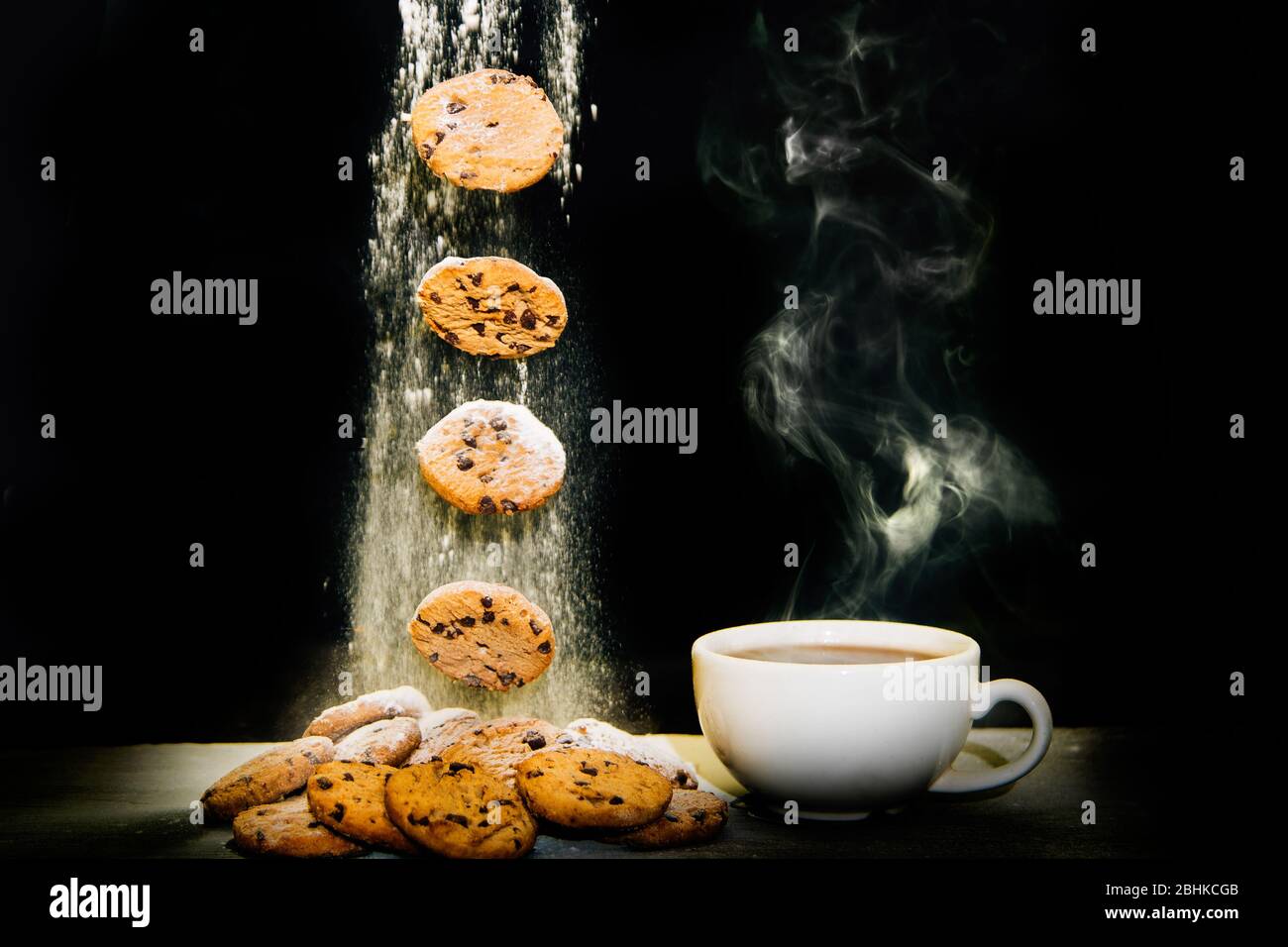 Thé et biscuits. Cookies ou biscuits tombant sur une pile de cookies à côté d'une tasse de thé à la vapeur sur fond noir. Farine saupoudrée de biscuits. Banque D'Images