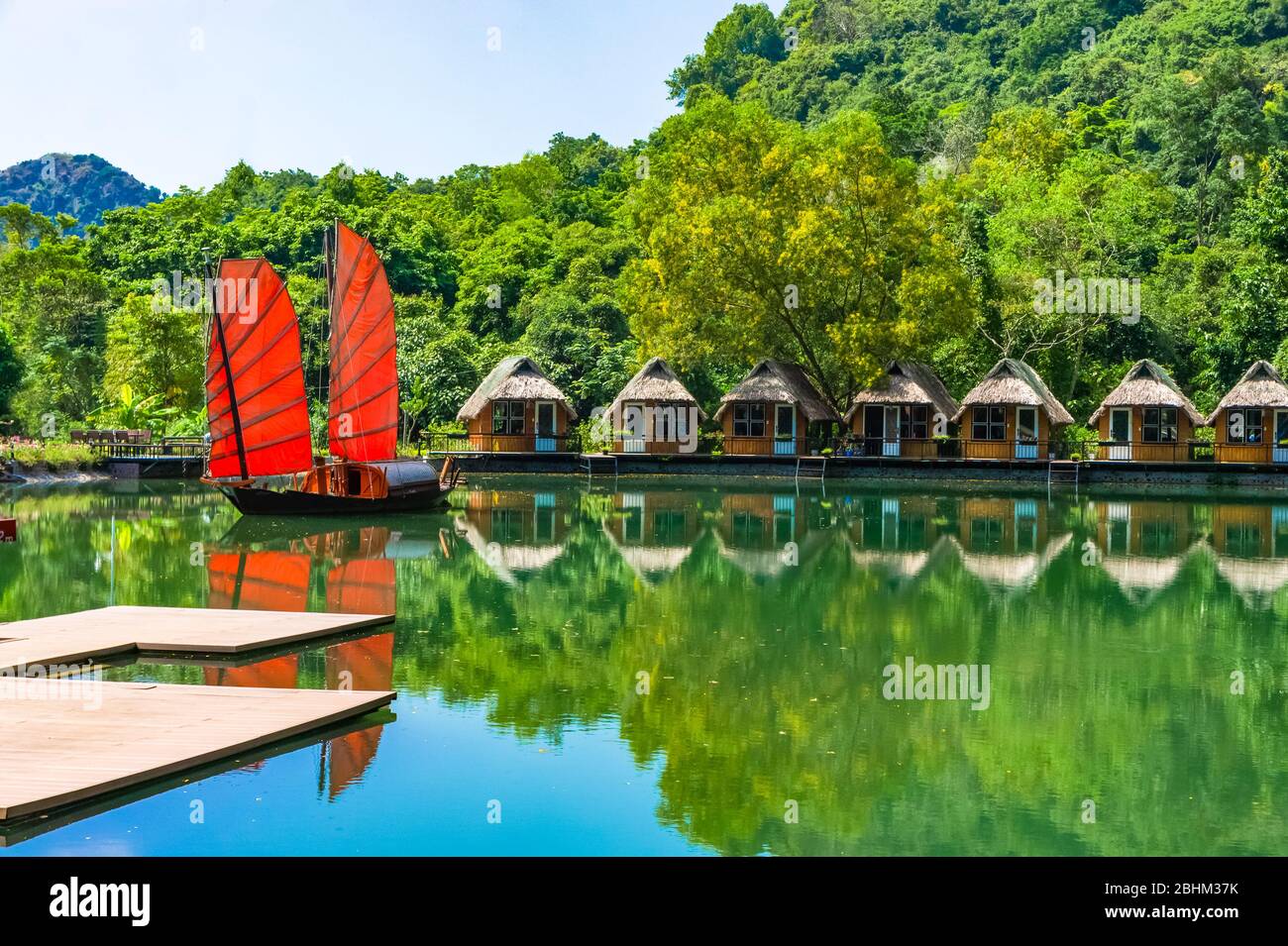 Indésirable. Magnifique bateau traditionnel vietnamien avec voiles rouges sur un lac pittoresque dans la jungle. De nombreuses petites maisons se reflètent dans l'eau. Banque D'Images