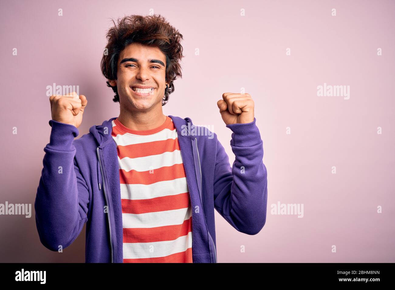 Jeune homme portant un t-shirt rayé et un sueur violet sur un fond rose isolé célébrant surprise et stupéfaite de succès avec les bras levés et Banque D'Images