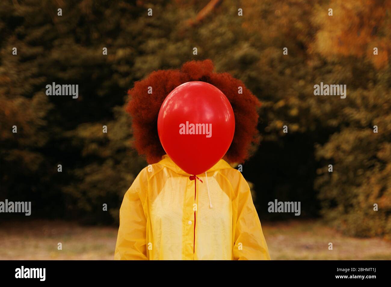 Une femme afro aux cheveux rouges dans un imperméable jaune tenant un ballon rouge sur le fond de la forêt. Concept Halloween, pas de visage Banque D'Images