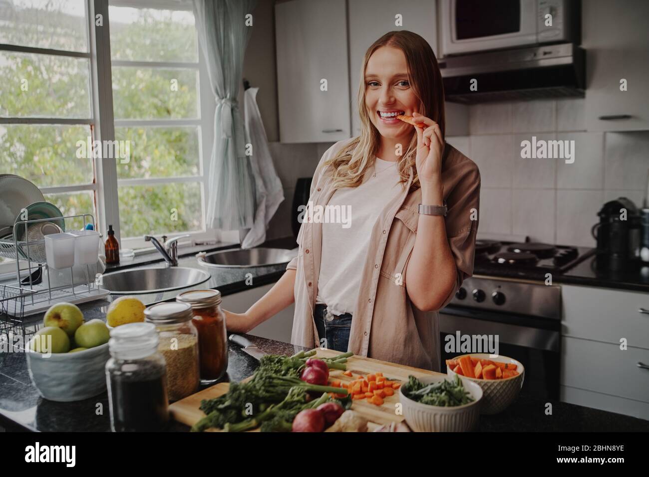 Jeune femme gaie dans la cuisine moderne préparant salade de légumes frais et manger une carotte souriant à l'appareil photo Banque D'Images