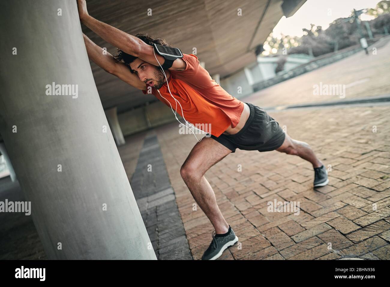 Portrait d'un homme de fitness avec brassard et écouteurs faisant des exercices d'étirement sous le pont Banque D'Images