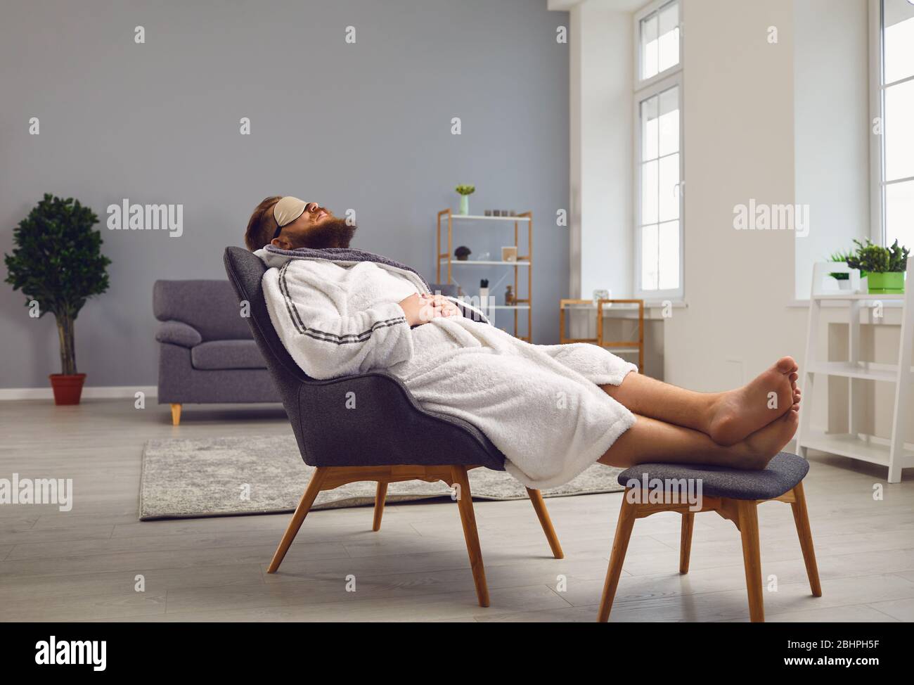 Un homme marrant barbu dans un peignoir dans un masque de sommeil se trouve  dans une chaise confortable dans le salon Photo Stock - Alamy