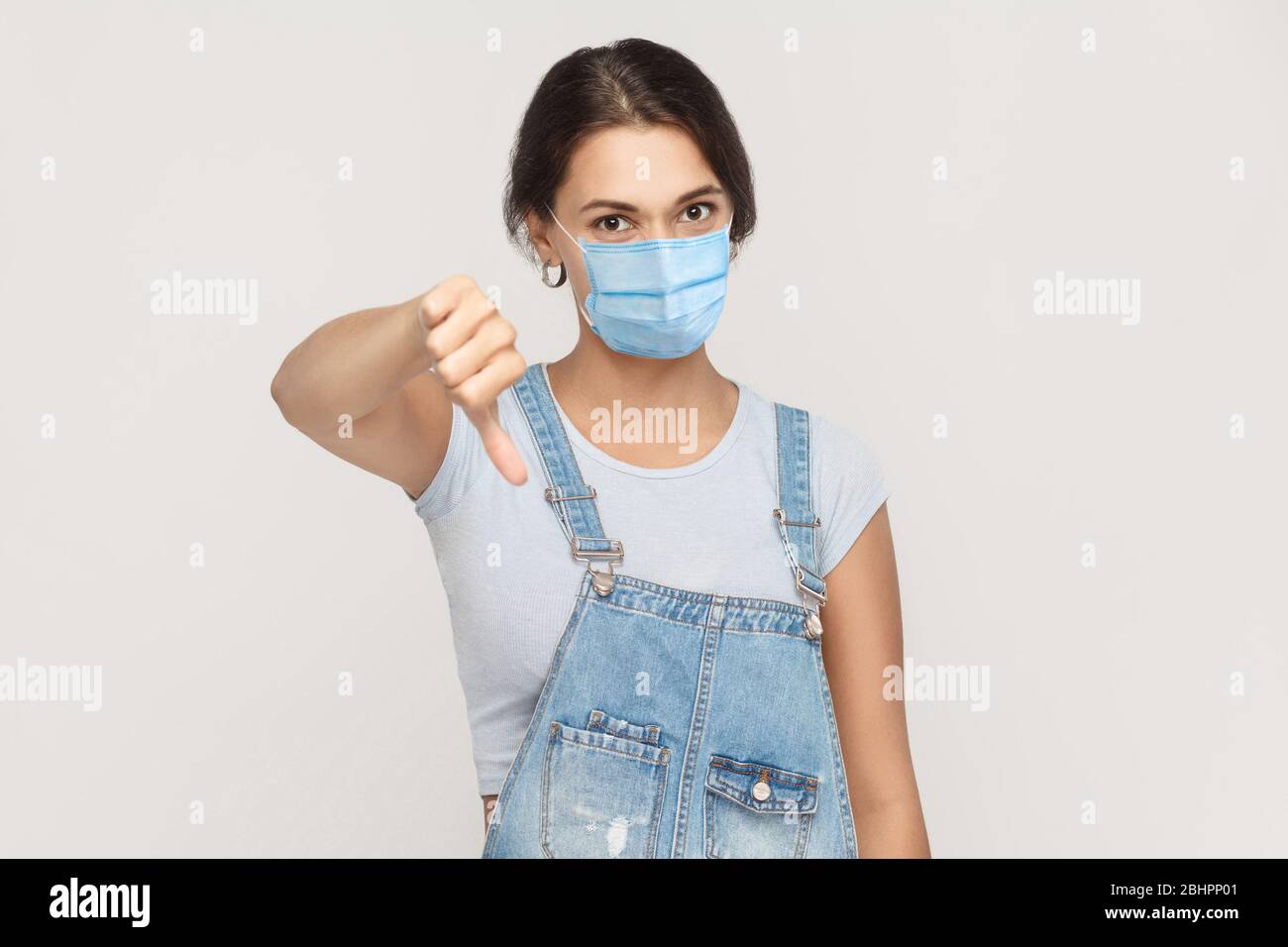 Je n’aime pas. Portrait de jeune femme mécontente de brunette avec masque médical chirurgical dans des salopettes en denim debout pouces vers le bas et regardant l'appareil photo Banque D'Images